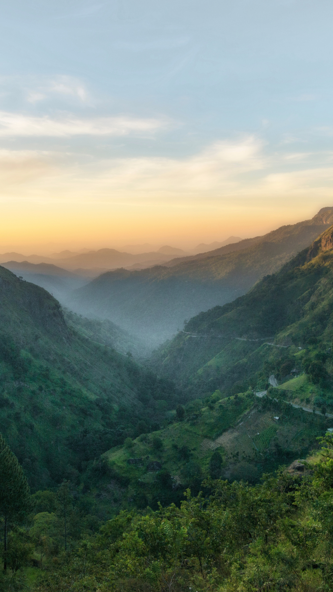 A scenic view of green mountains and valleys during sunrise or sunset, with a cloudy sky and mist in the distance.