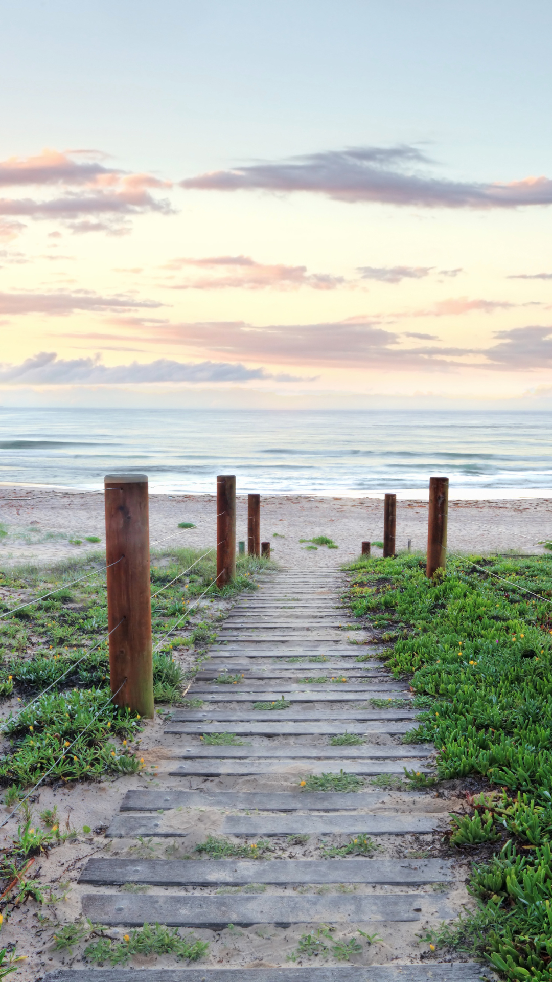 Wooden pathway leading to a beach with sand and ocean, surrounded by green vegetation under a cloudy sky.