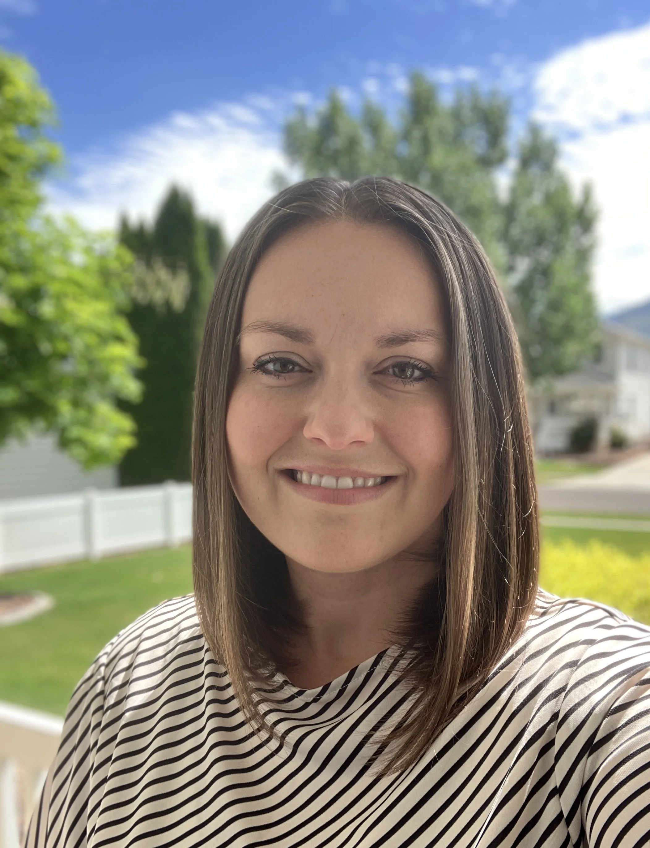 Close-up of a smiling woman with long brown hair, wearing a striped shirt, outdoors with green trees and a white fence in the background on a sunny day.