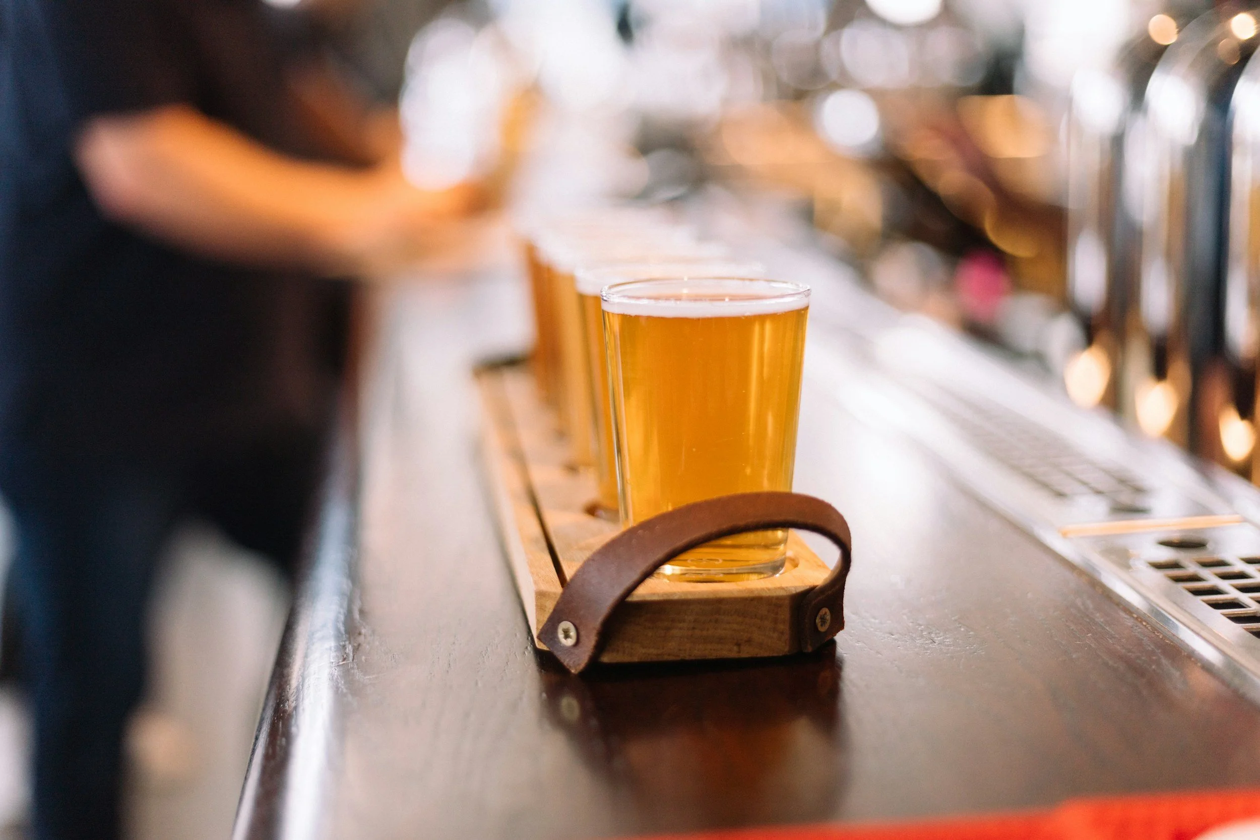 A glass of beer on a wooden tray in a bar or pub setting, with a row of other beers on tap in the background.