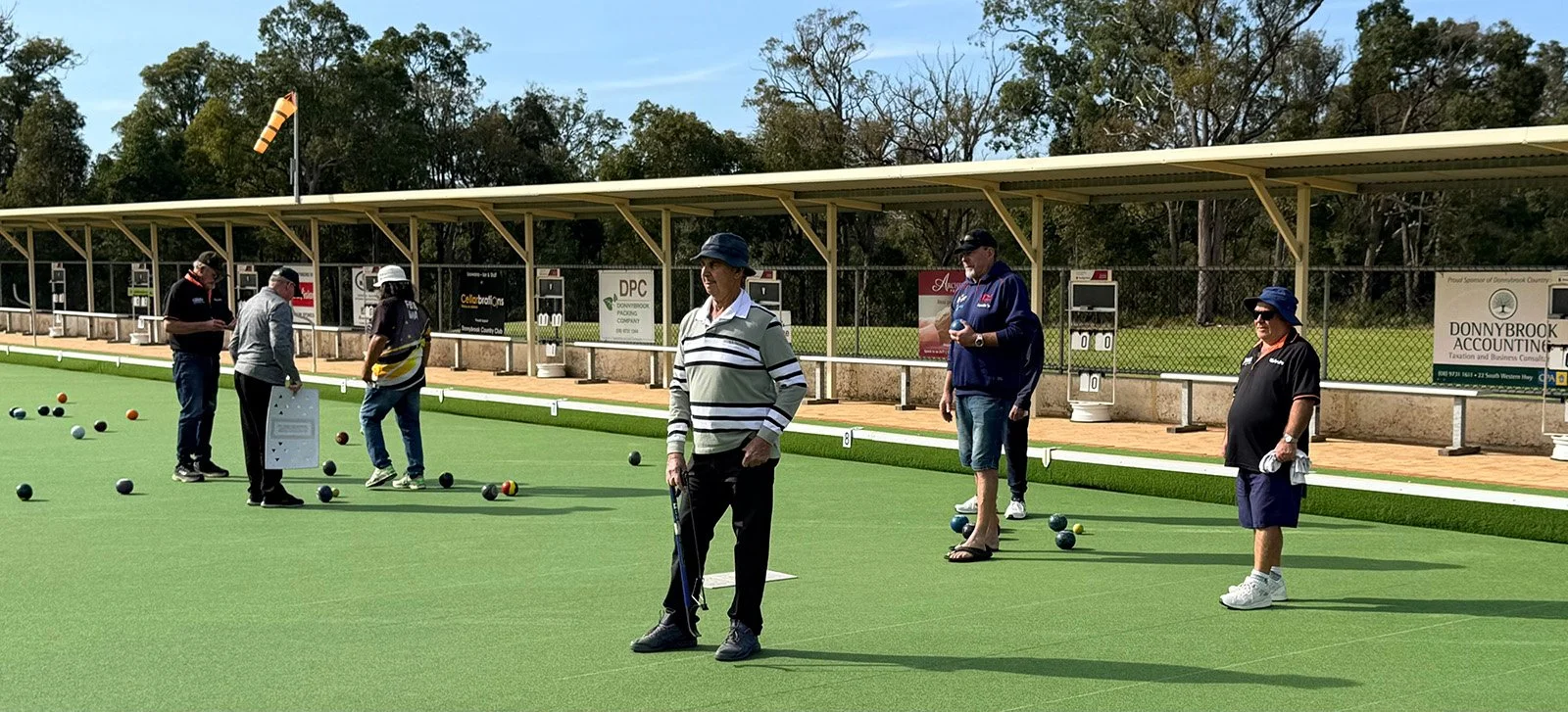 People playing lawn bowls on a green in Donnybrook, with some holding bowls and others observing, under a clear sky.