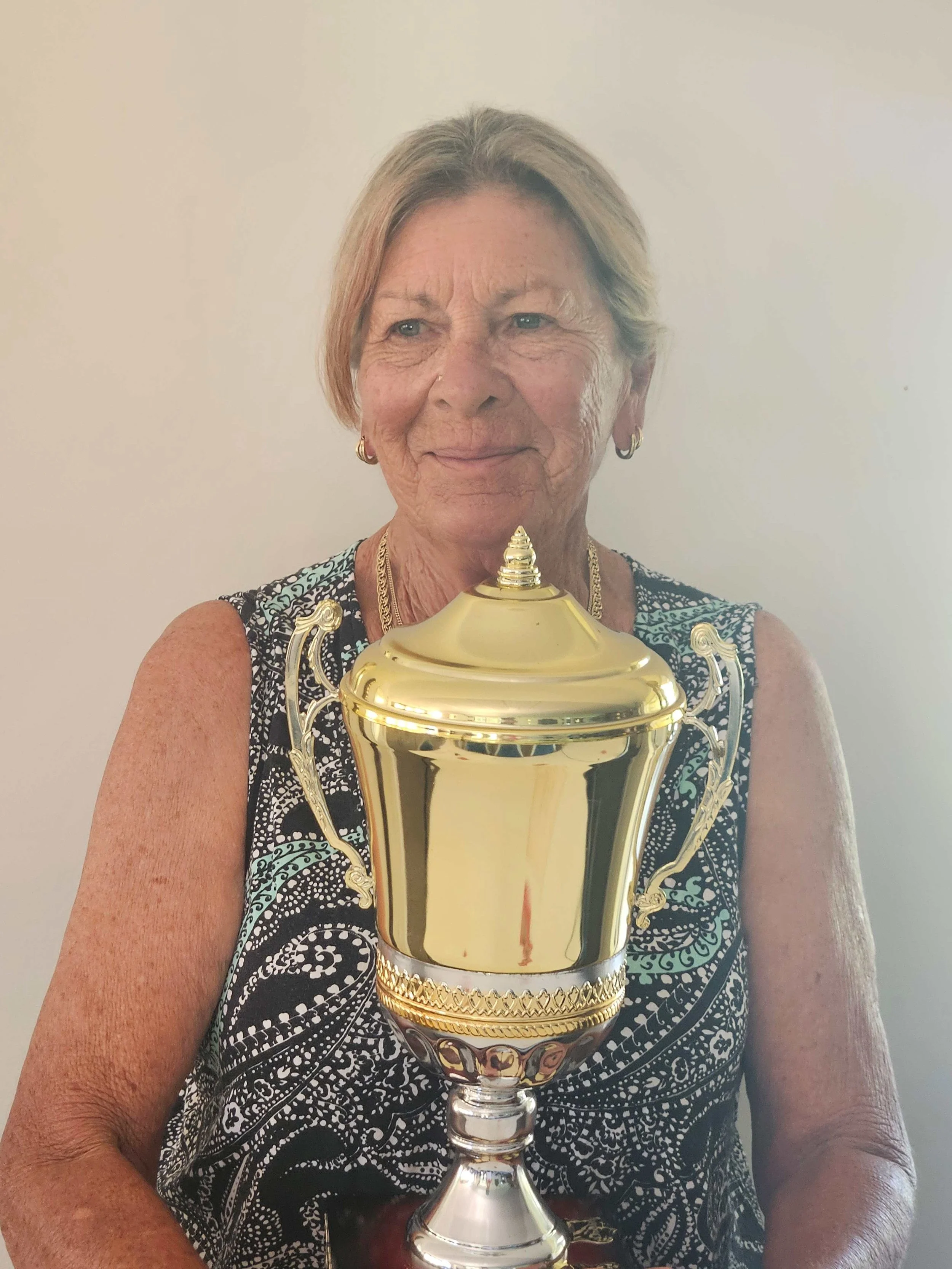 An elderly woman with short, light-colored hair holding a large, gold trophy in front of a plain wall.