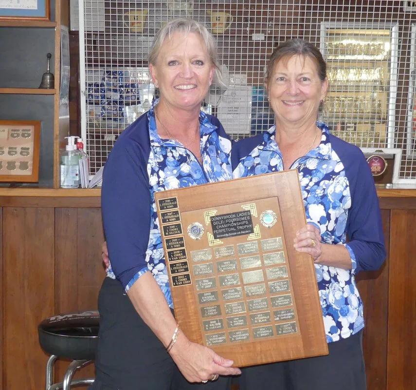 Two women in matching blue floral shirts holding a wooden plaque with multiple small plaques, smiling, standing in front of a wooden counter and wire mesh display, with a bottle of hand sanitizer visible.