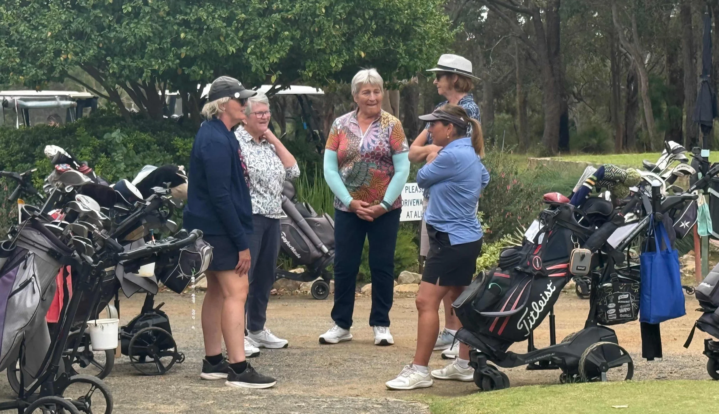 Group of women in golf attire standing near golf carts and golf bags outdoors, engaged in conversation.