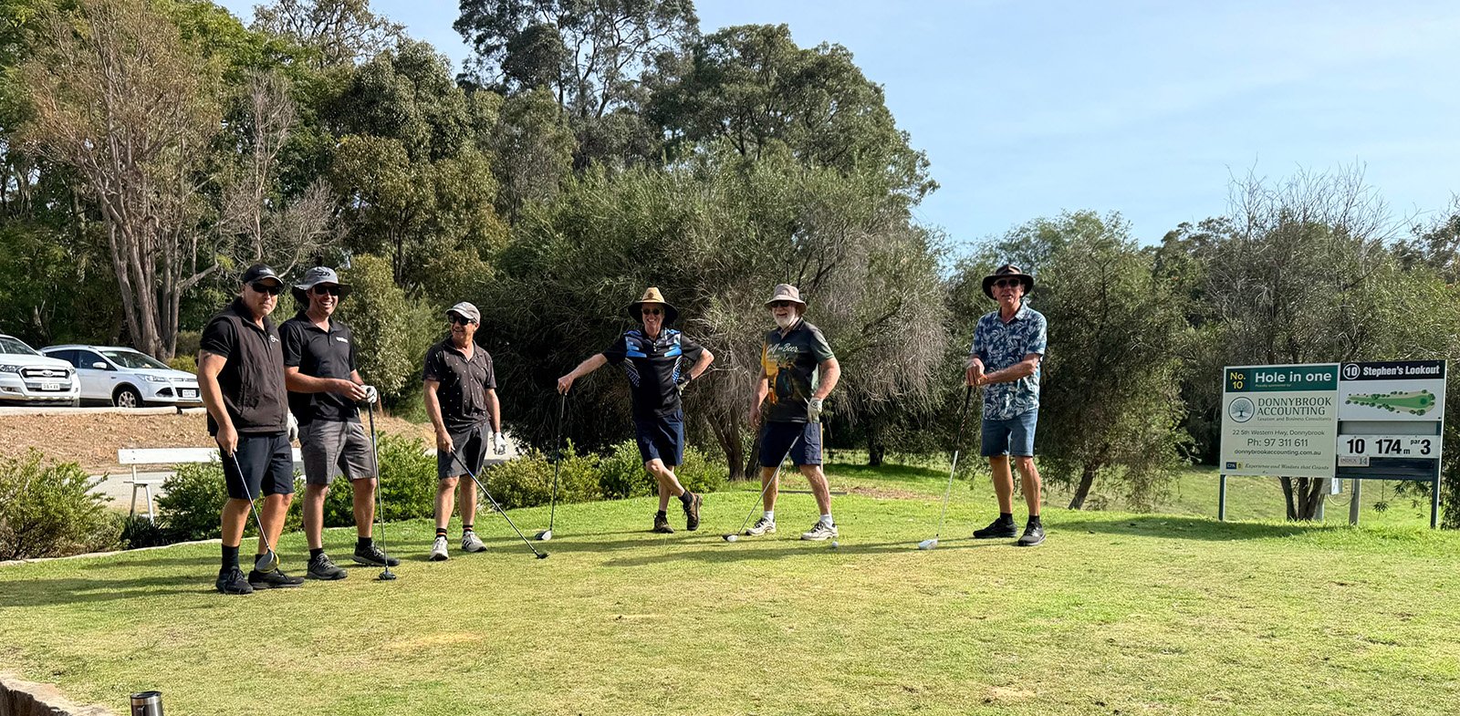 Six men standing on Donnybrook golf course, holding golf clubs, preparing to tee off. There are trees in the background, a parking lot with cars on the left, and a sign on the right indicating hole number 10 at a golf course.
