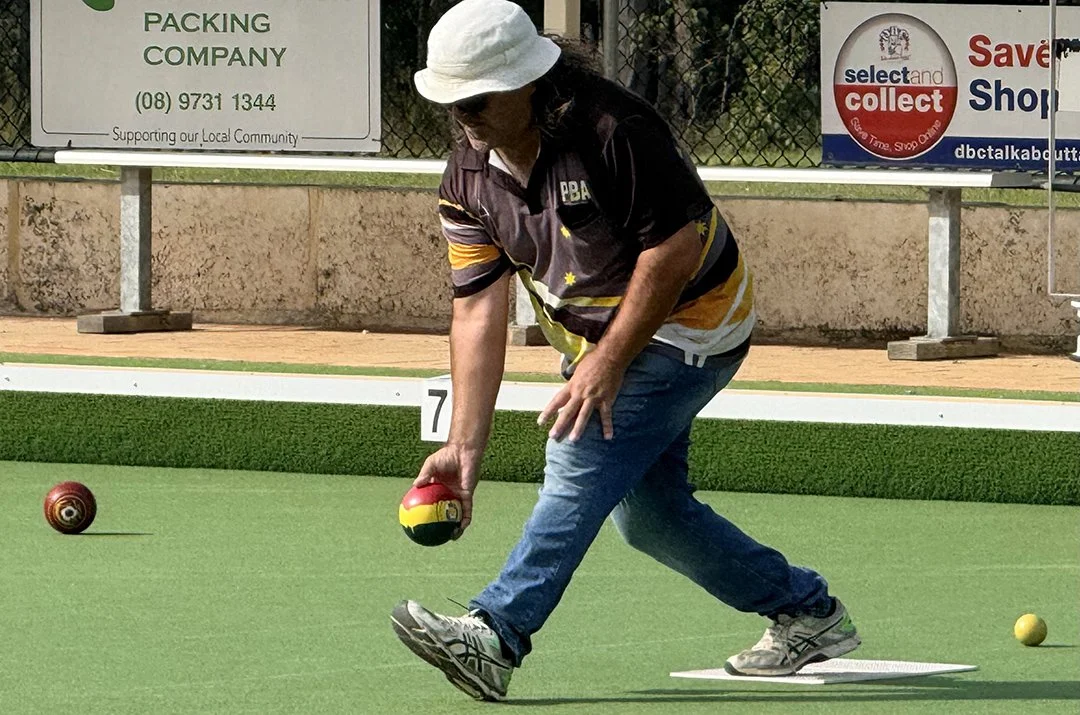 Man in a black polo shirt and light blue jeans playing lawn bowls on a green outdoor court in Donnybrook, holding a red, yellow, and black bowling ball.