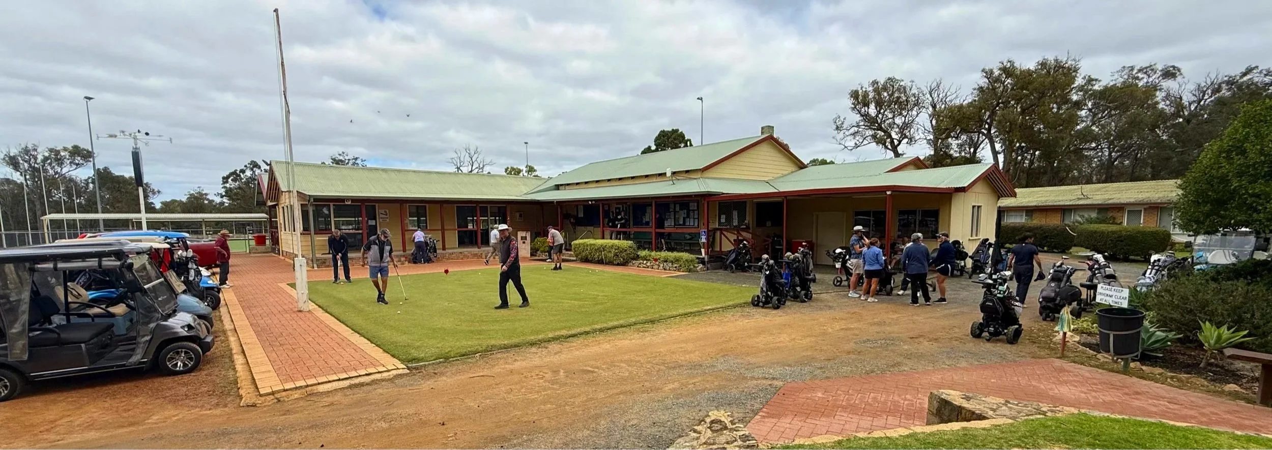 Donnybrook golf clubhouse with people preparing to play golf, some on the putting green and others near golf carts, under a cloudy sky.