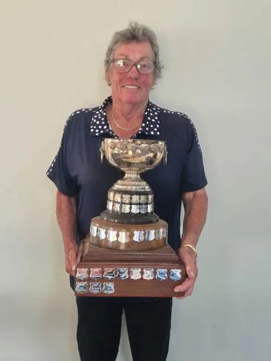An elderly woman holding a large trophy with a wide bowl and ornate base, standing against a plain light-colored wall.