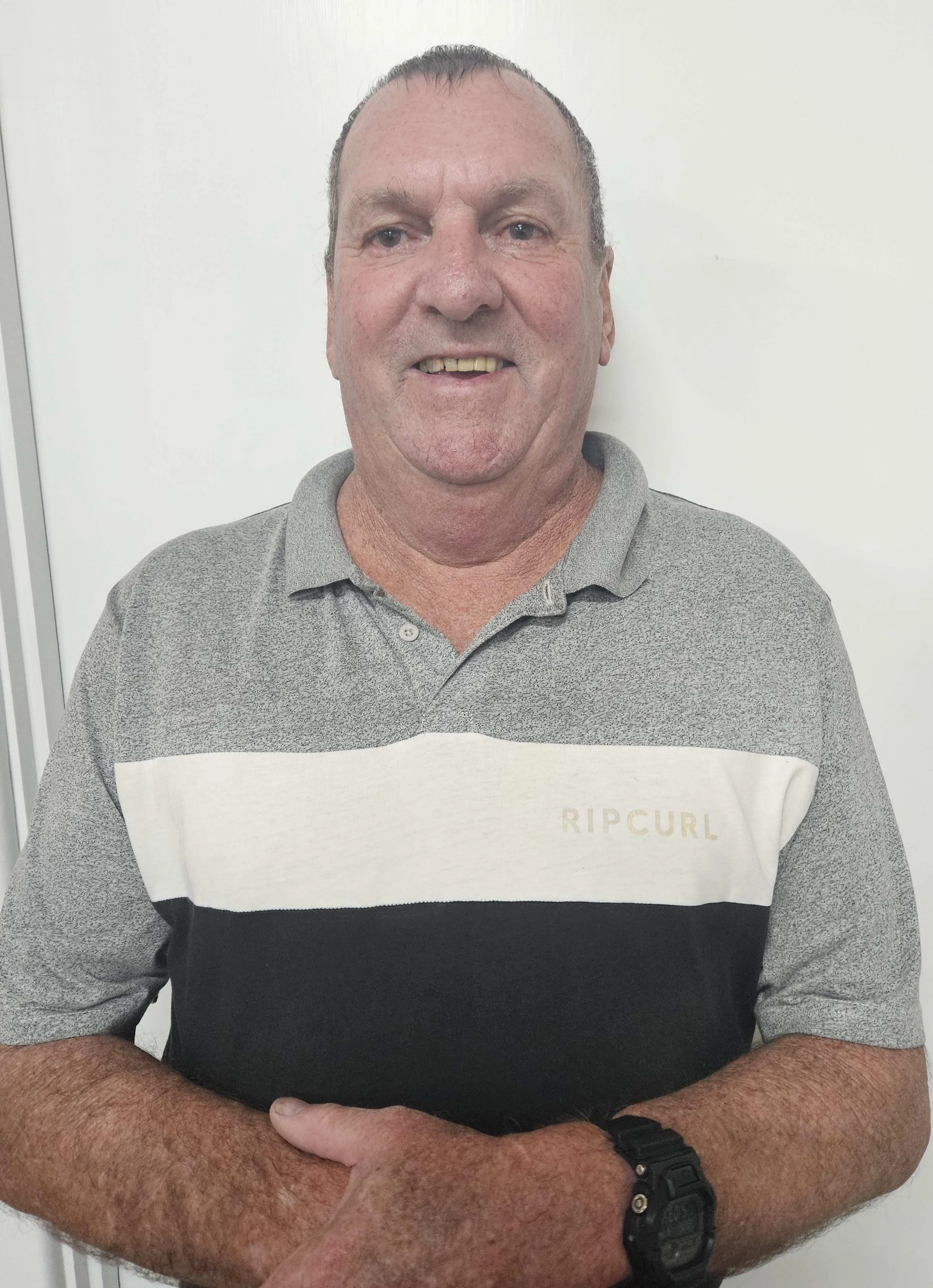 A middle-aged man with short, dark hair, wearing a gray Rip Curl polo shirt with a black and white horizontal stripe, standing with crossed arms in front of a plain white background.