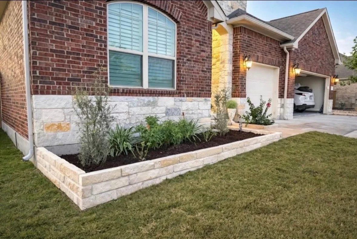 Front yard with raised flower bed of brick and stone near a brick house with large window and attached garage, with a white car inside, and outdoor wall lights.