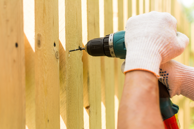Person wearing gloves using a power drill to drill into wooden fence panels.