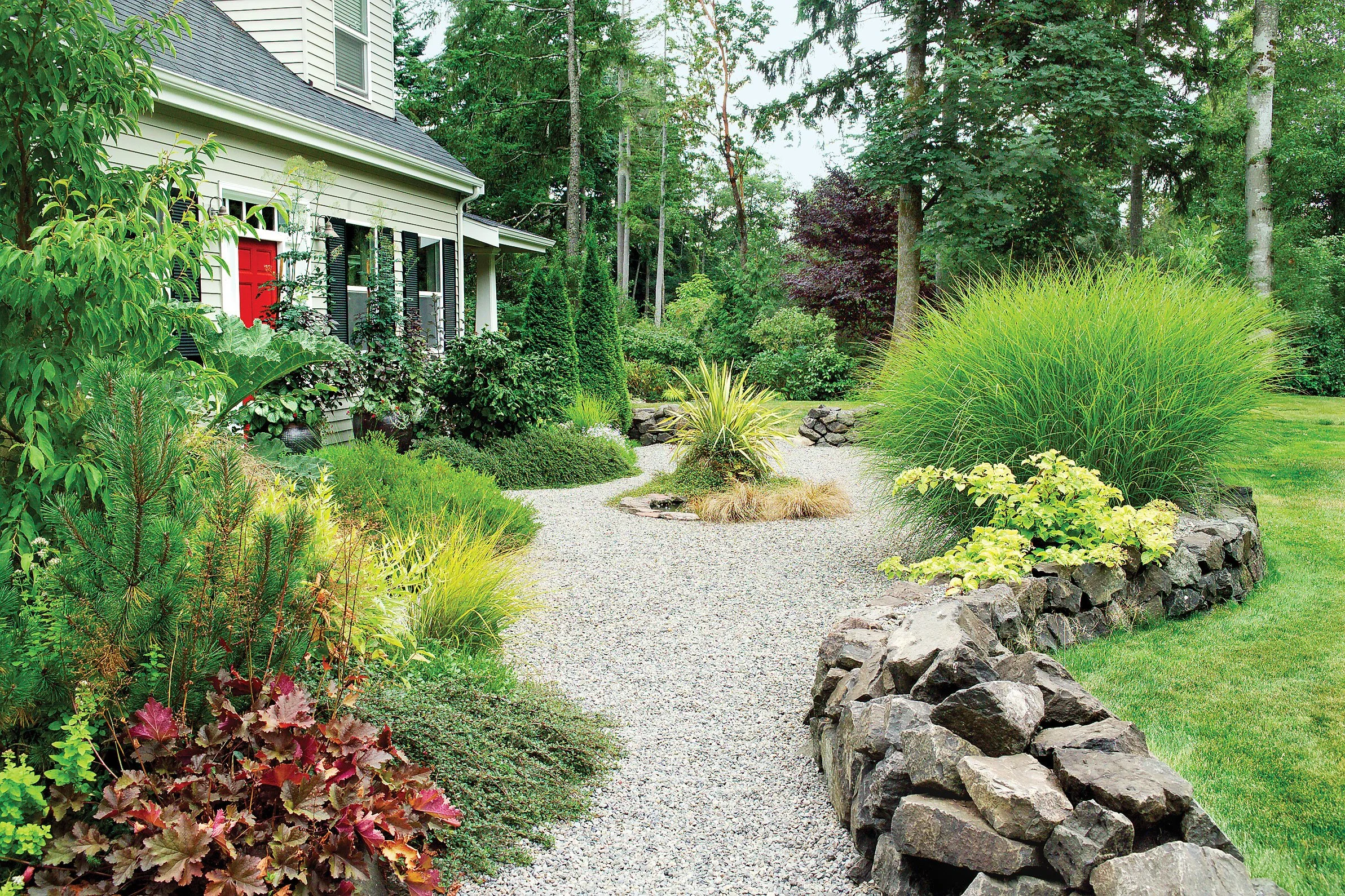 A well-maintained gravel garden path lined with lush green plants and trees, next to a house with white siding and black shutters.