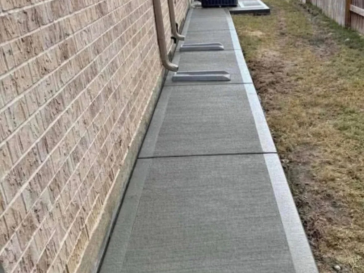 A sidewalk with metal wheelchair ramps along a brick building and a grassy area on the side, with a trash bin in the background.
