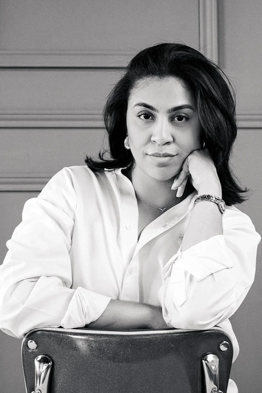 Black and white photo of woman with dark hair sitting behind a chair, resting her chin on her hand, wearing a white shirt.