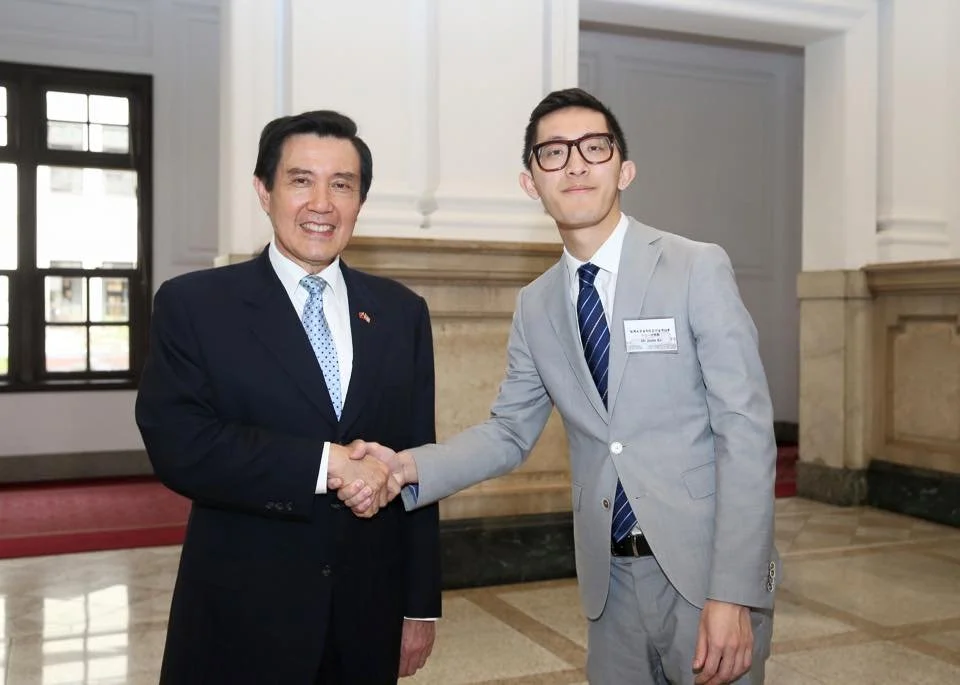 Two men in suits shaking hands indoors, smiling, with architecture featuring large windows and marble details in the background.