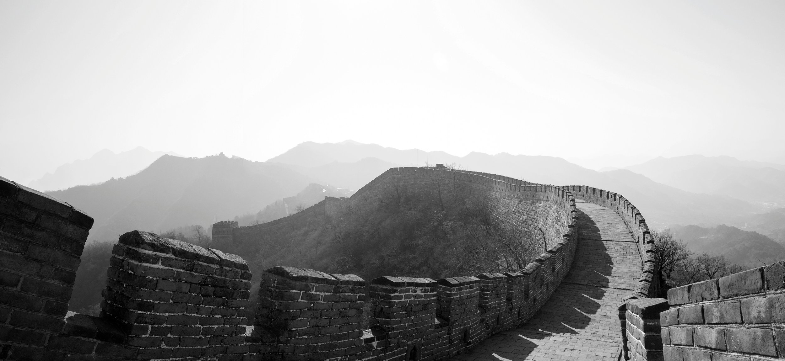 Black and white photo of the Great Wall of China winding through mountainous terrain with distant mountain peaks in the background.