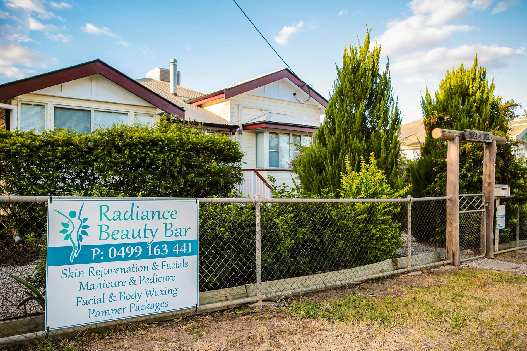House surrounded by trees and bushes, with a fence and sign for Radiance Beauty Bar in front, advertising skin treatments, manicures, pedicures, facials, waxing, and packages, under a blue sky with clouds.