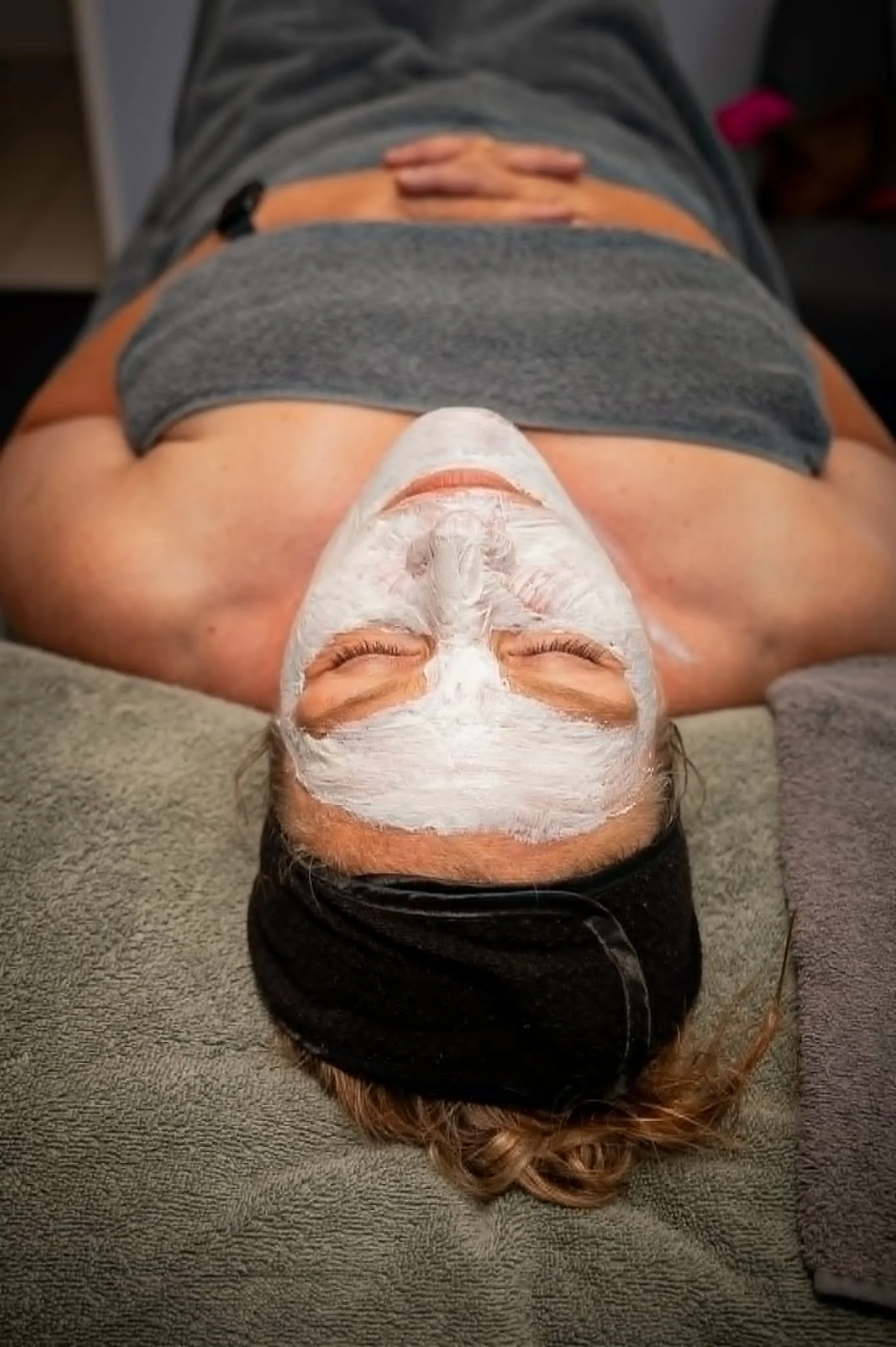A person lying on a spa table with eyes closed, wearing a black headband and a grey towel, having a white facial mask applied.