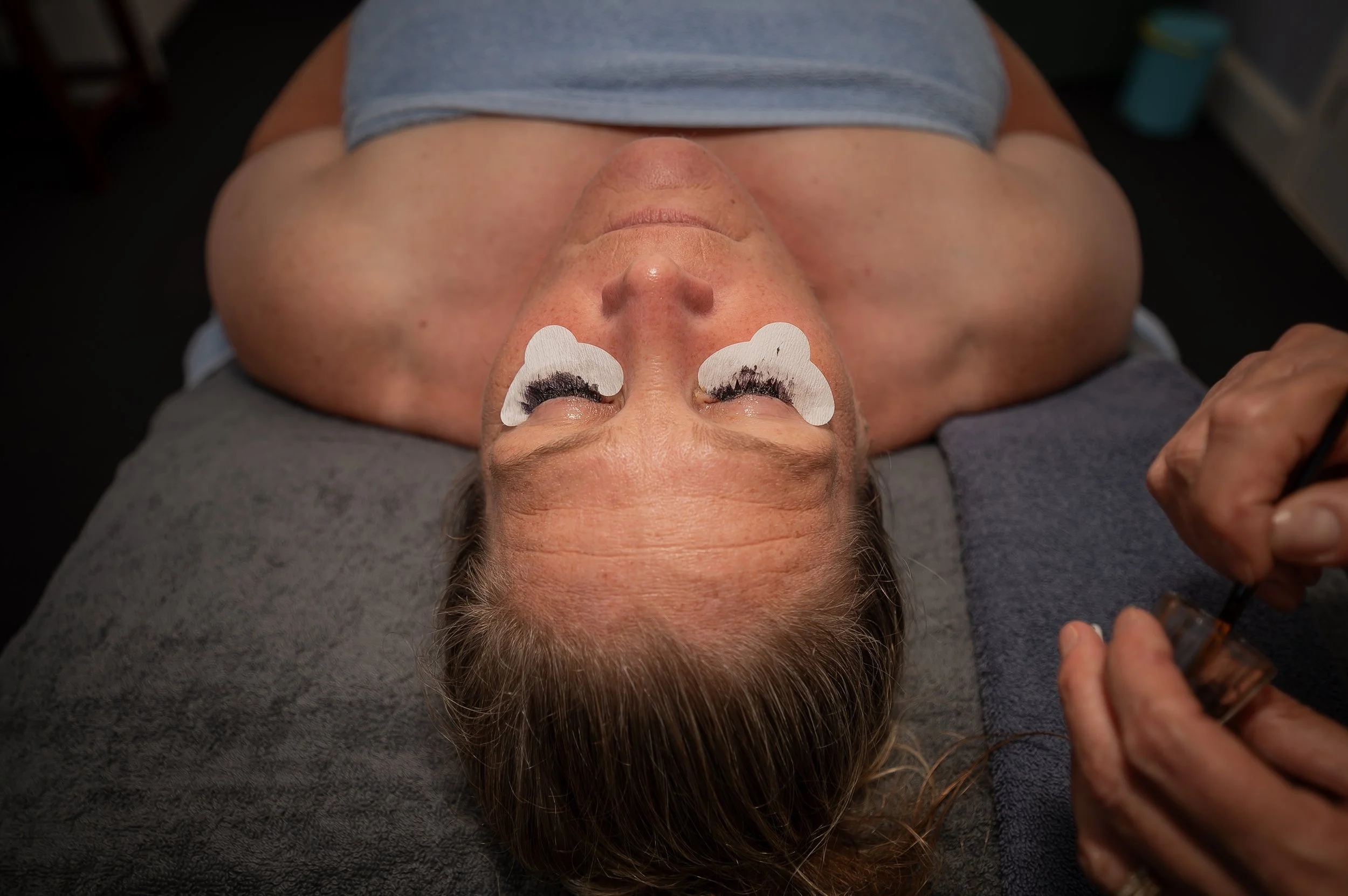 A woman lying on a treatment table with eye patches and eyelash extensions being applied by a technician.