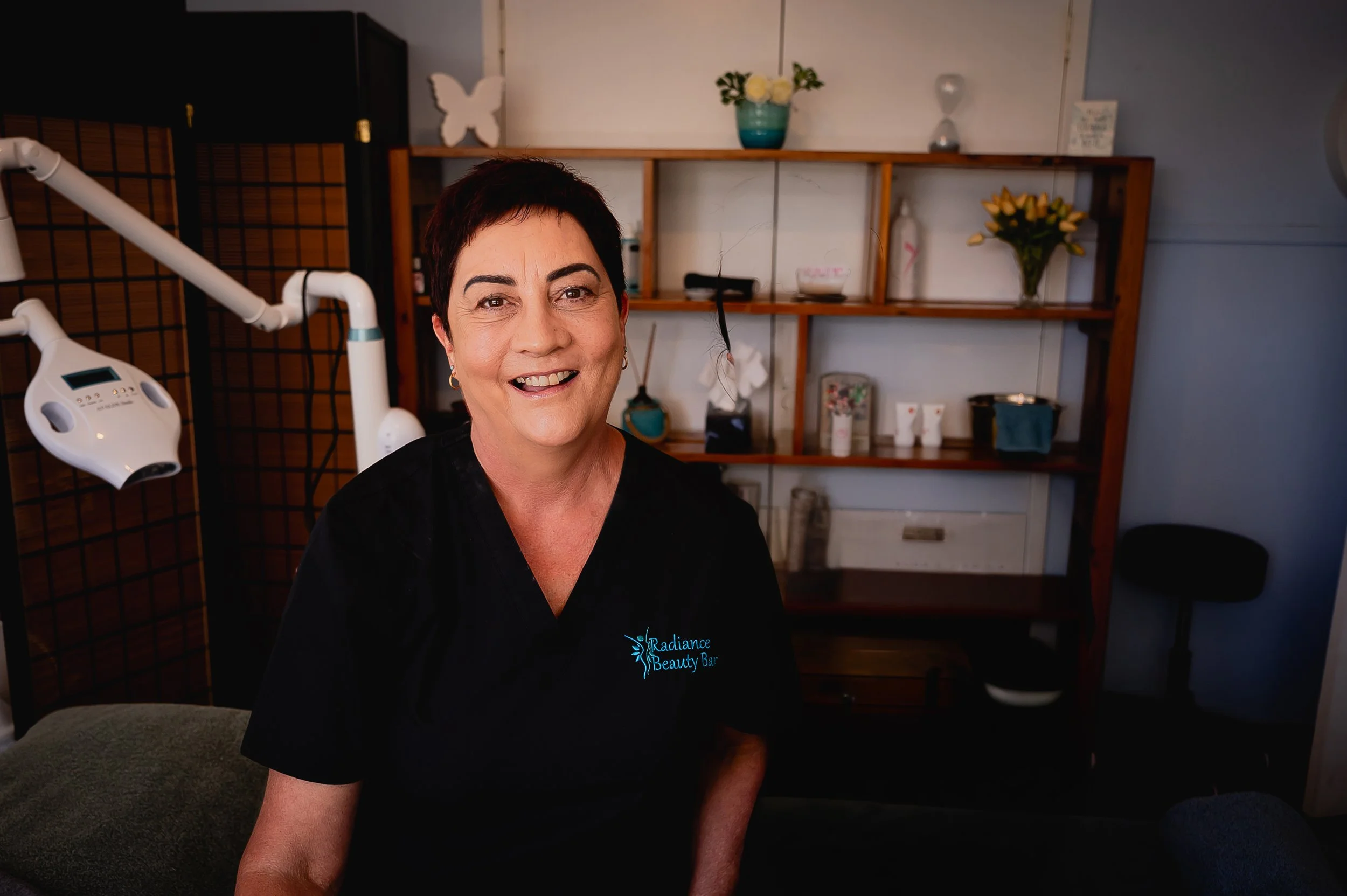 A smiling woman with short dark hair wearing a black shirt with the Radiance Beauty Bar logo, standing in a room with shelves and decorative items in the background.