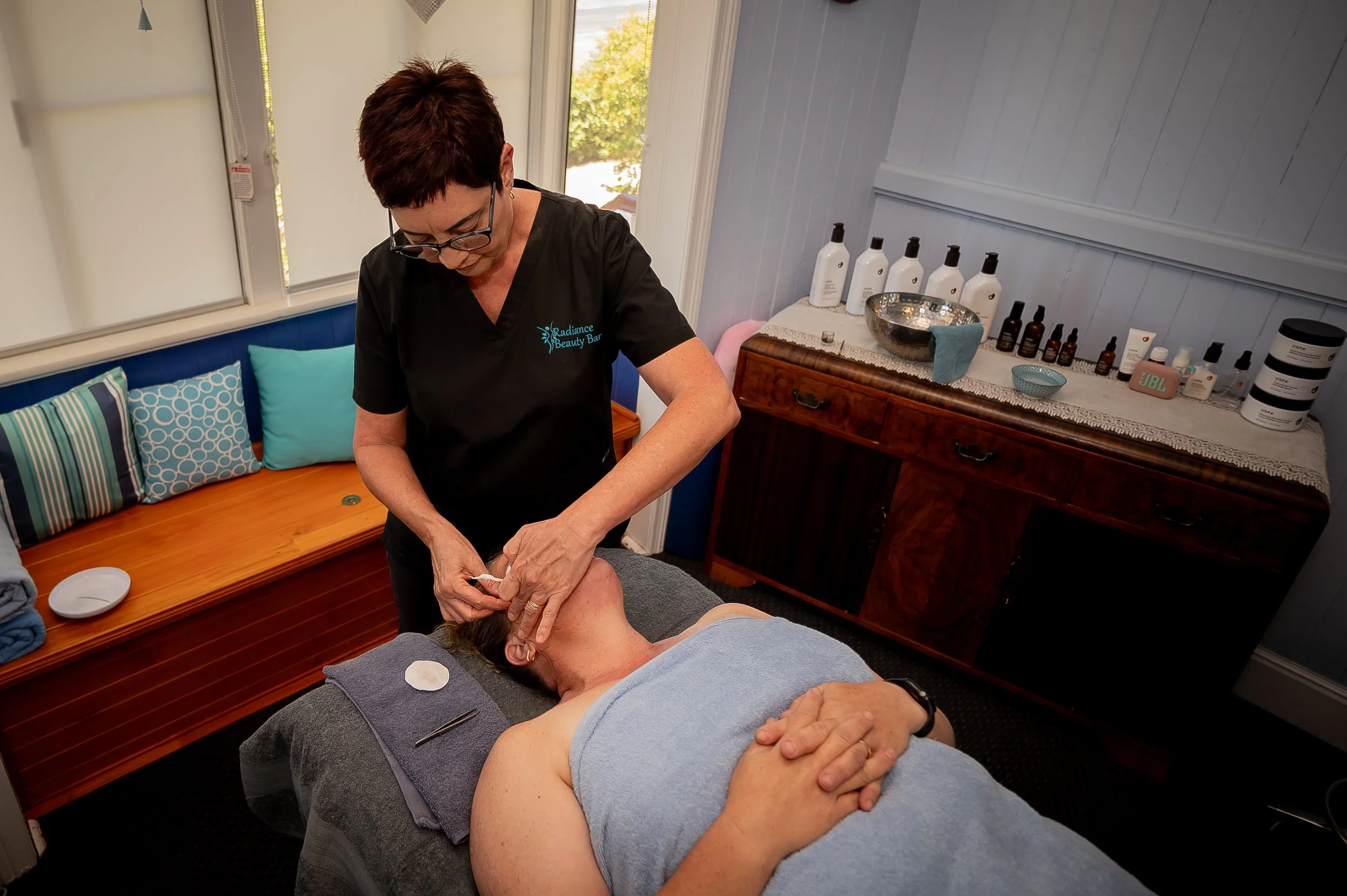 A woman receiving a facial treatment in a spa or beauty salon, lying on a treatment bed with her eyes closed, while a professional offers a skincare procedure. The room is decorated with colorful cushions on a bench, and various skincare products are