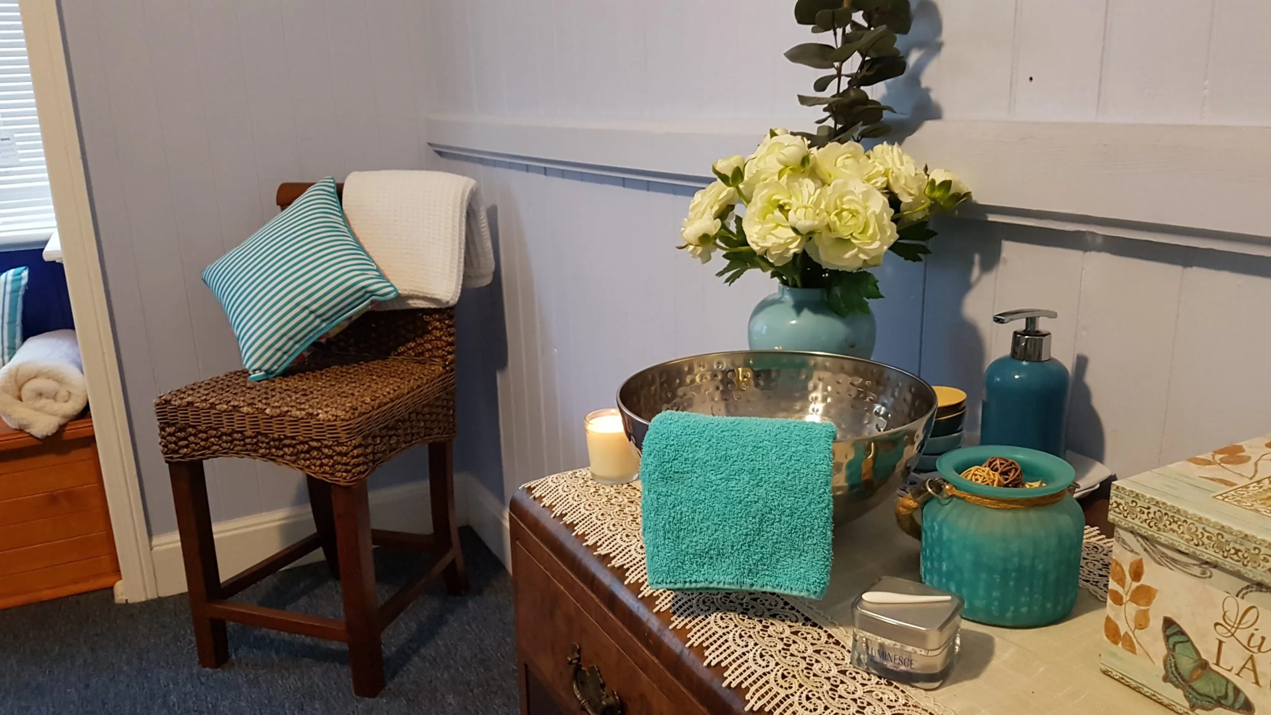 Decorative table with a large vase of white flowers, a turquoise towel, a candle, a metallic bowl, teal soap dispenser, smaller teal containers, a woven basket, and a box with floral design in a cozy room with a wicker chair and a cushion.