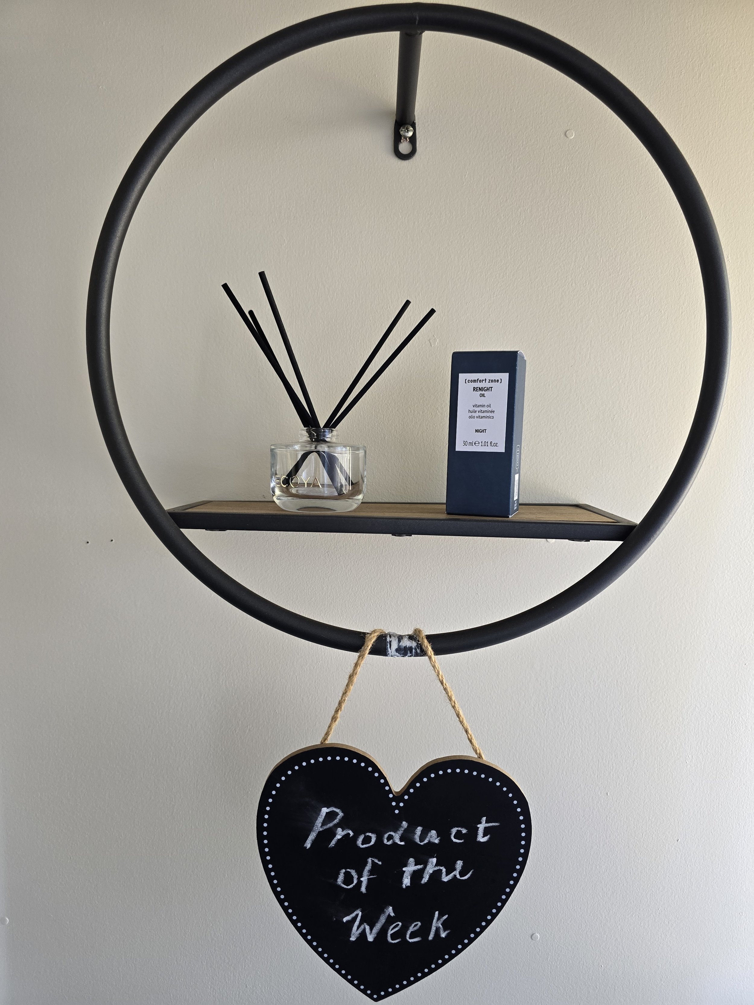 A wall clock with a black frame and black hands. Below the clock, a small glass bowl holds black reed diffuser sticks. To the right, a blue box of night oil is visible. A heart-shaped chalkboard sign hanging below says, "Product of the Week."