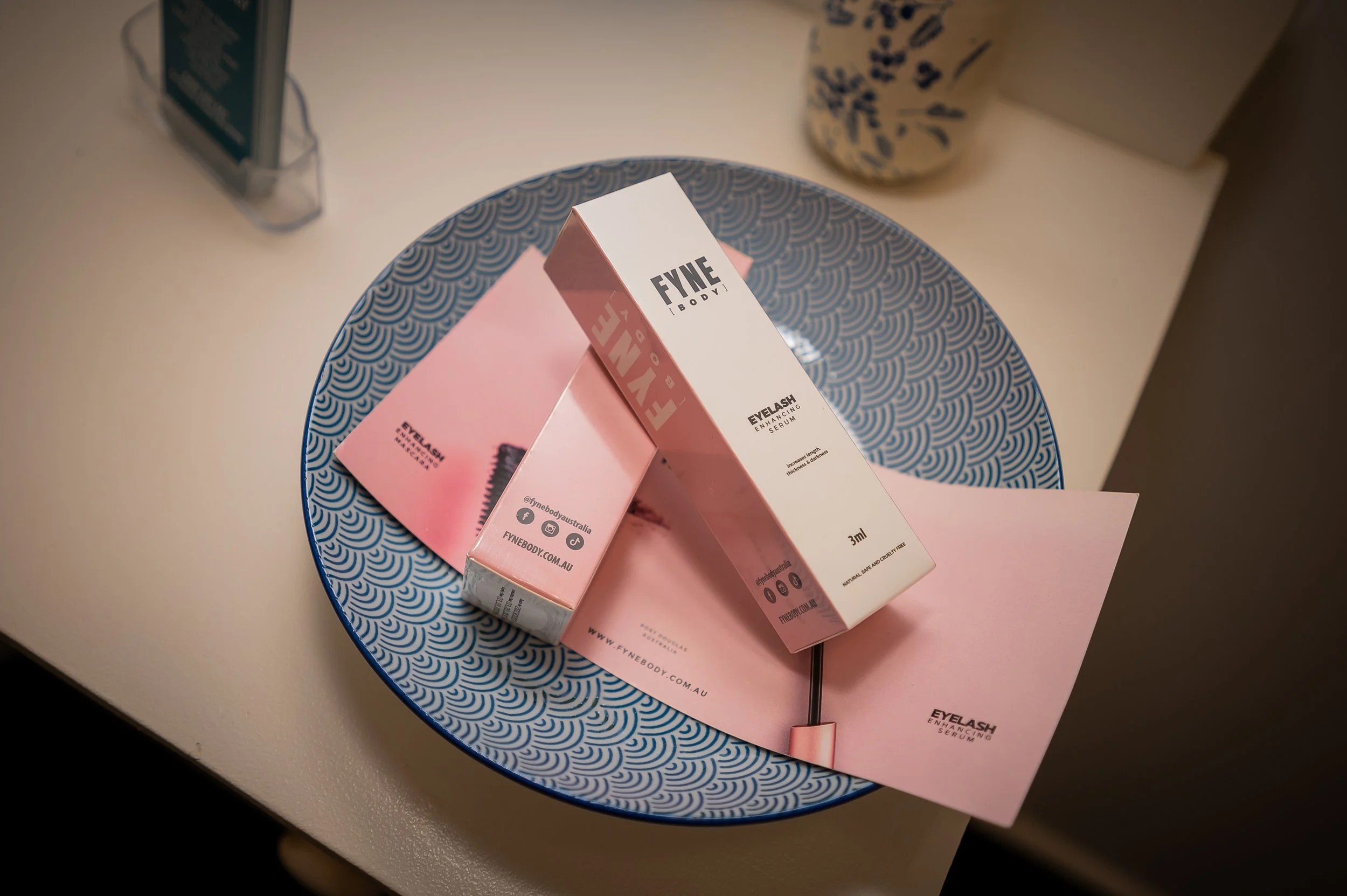 A pink box labeled FYNÉ EYELASH ENHANCING SERUM placed on a blue and white patterned round table.