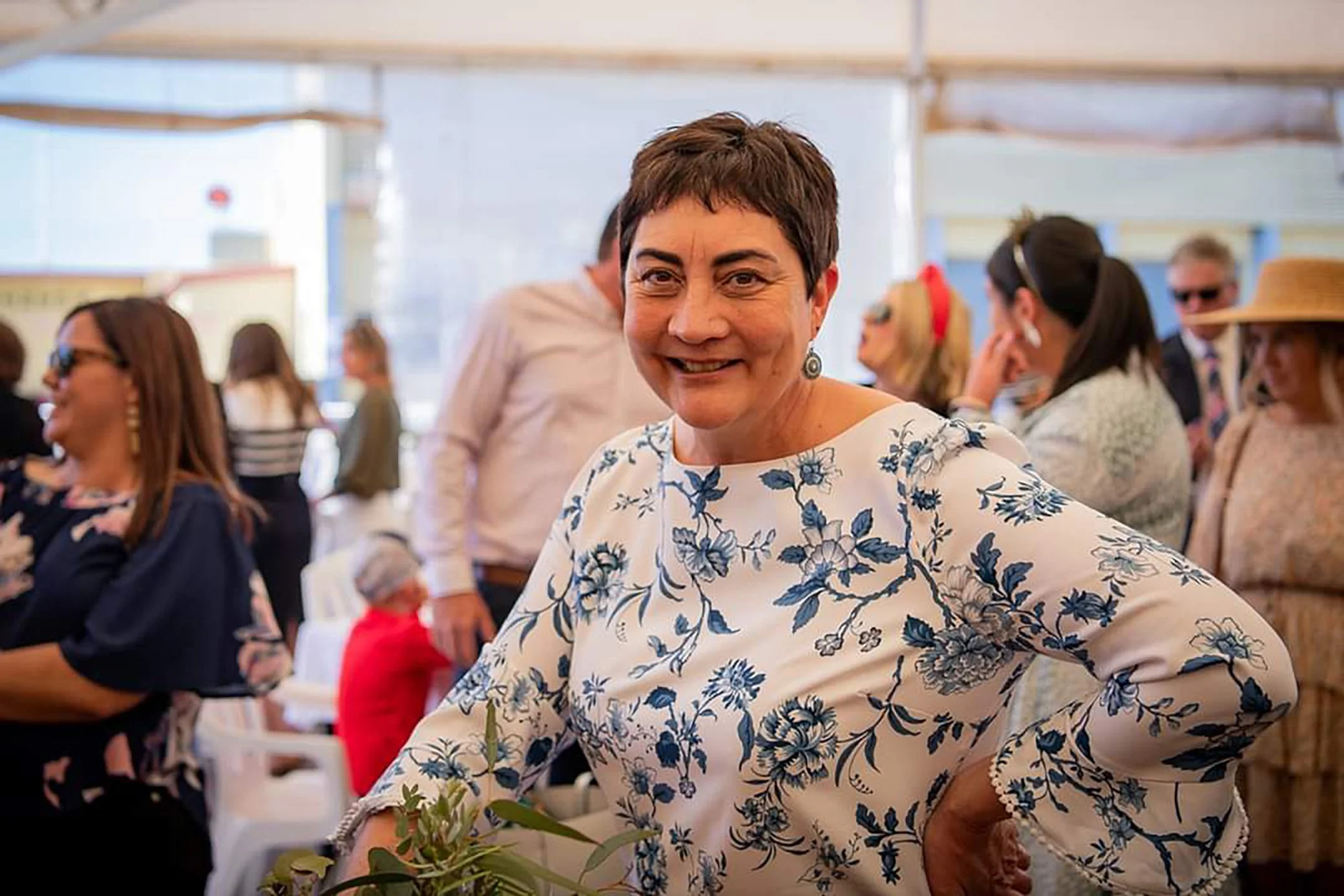 A woman with short brown hair, smiling and wearing a white dress with blue floral patterns, at a social event. Other people are in the background, some wearing sunglasses and casual clothing, inside a bright space with large windows.