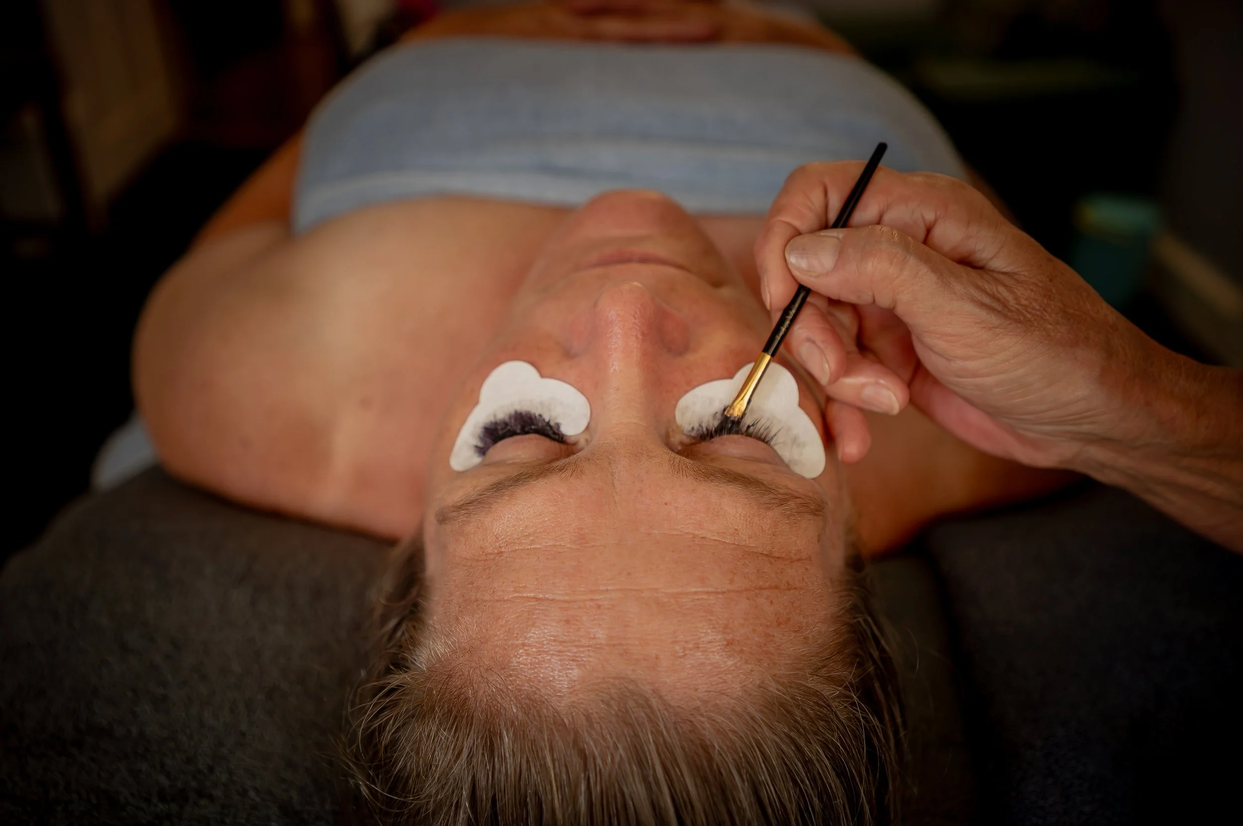 A person lying down with eyes closed, receiving a facial treatment or facial massage, with cotton pads under their eyes and a hand holding a brush or applicator near their face.