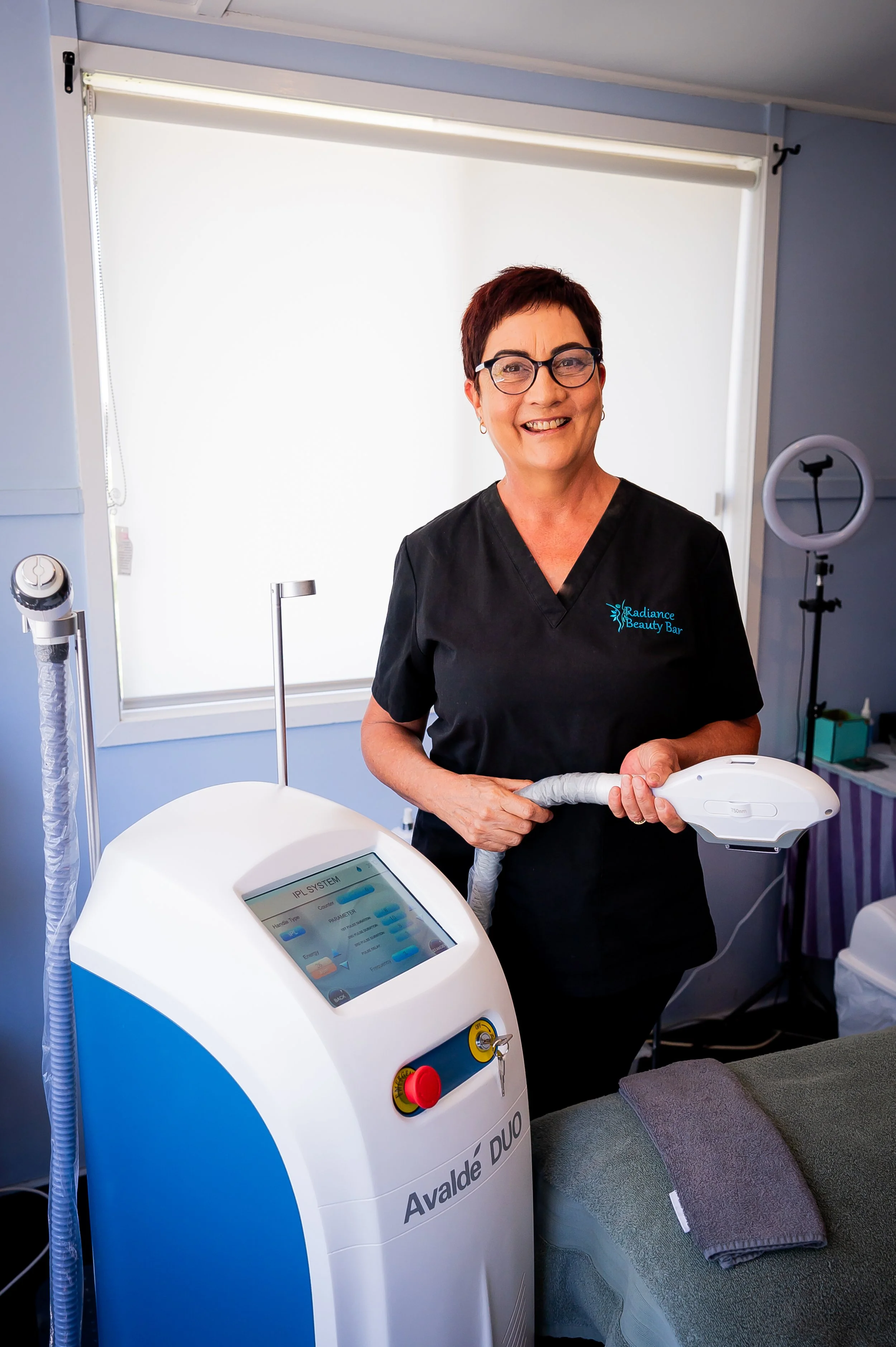 A woman with short red hair, glasses, and wearing a black medical uniform with the logo 'Radiance Beauty Bar', smiling while holding a handheld device in a medical or beauty treatment room. There is a machine labeled 'Avalde DUO' with a touchscreen, 