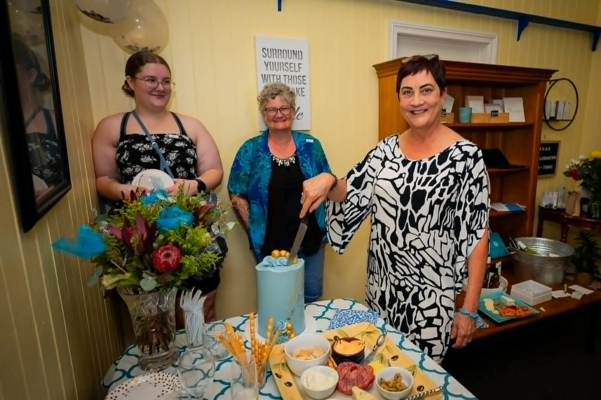 Three women smiling and exchanging a handshake at a celebration or gathering, with tables of food and flowers in the foreground and a yellow wall with a sign that says 'Surround yourself with those who make you smile' in the background.