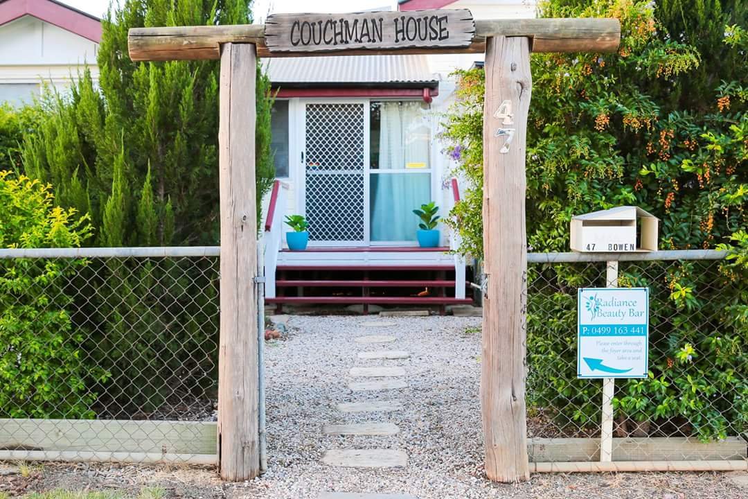 Wooden gate with a sign that reads 'Couchman House' leading to a house with a screened door, potted plants, and steps, surrounded by green bushes and trees.