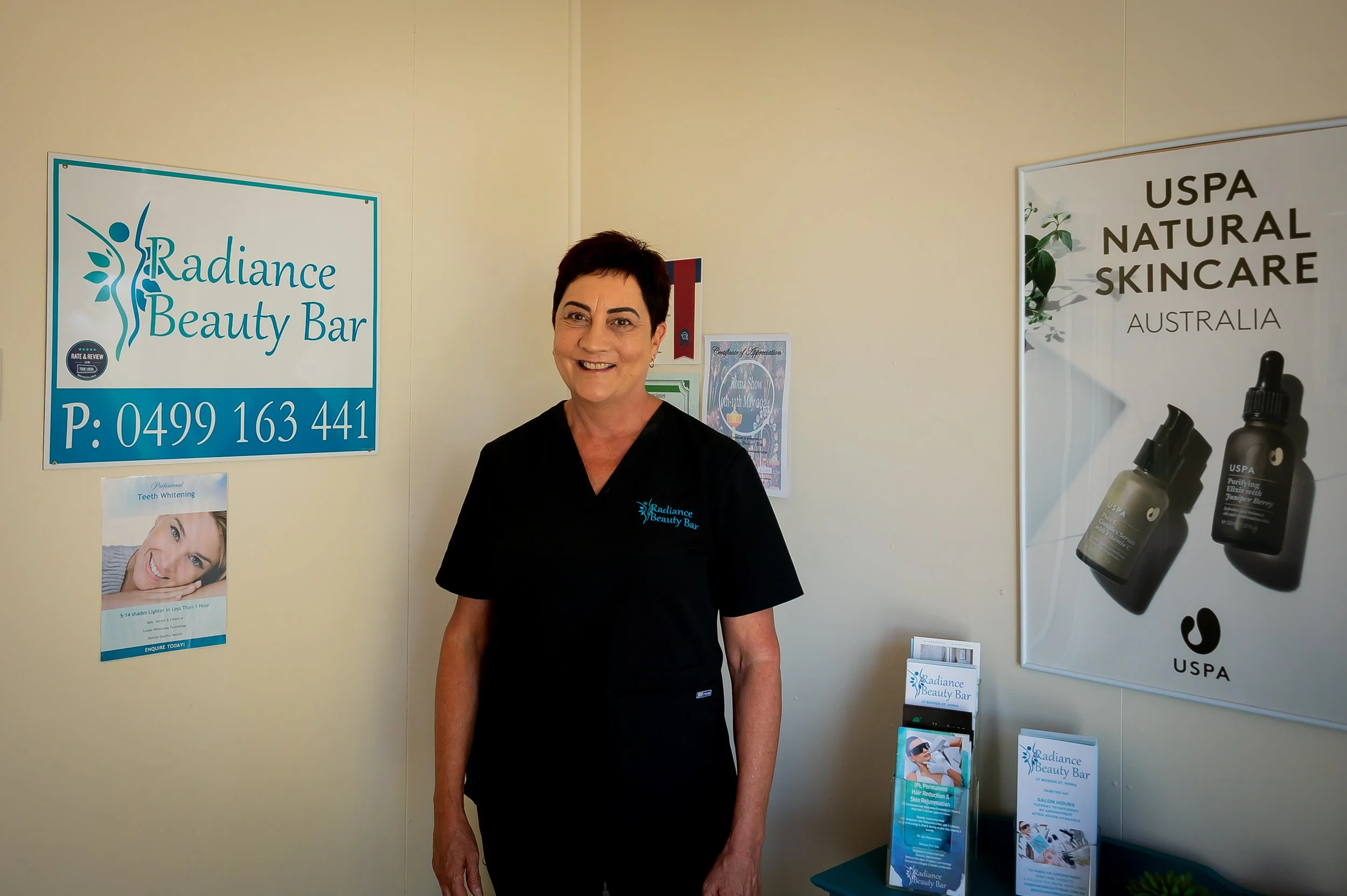 A woman with short dark hair and wearing a black shirt standing indoors near walls with signs for Radiance Beauty Bar and USDA Natural Skincare, smiling at the camera.