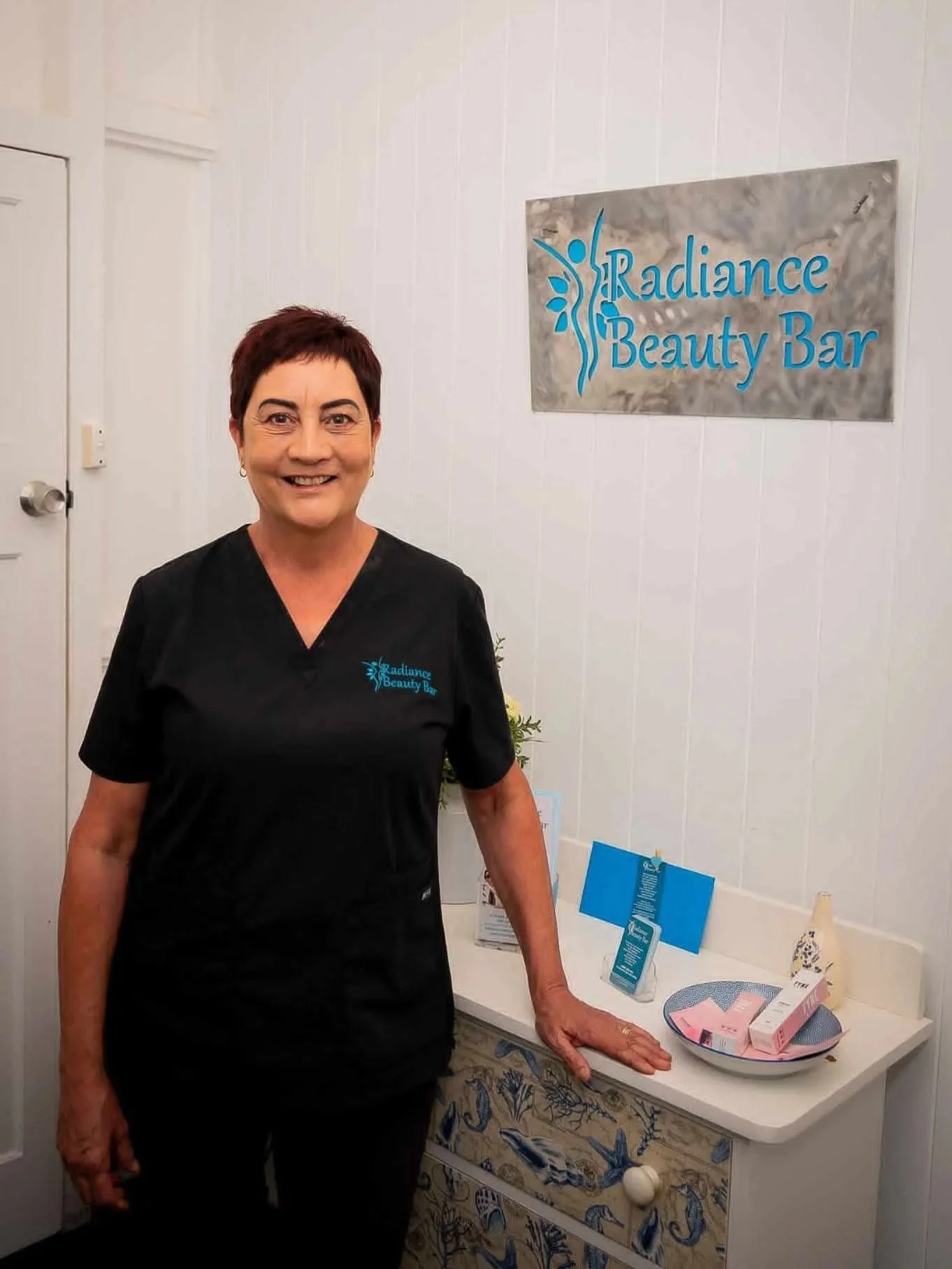 A smiling woman wearing a black uniform standing inside a beauty or wellness clinic called Radiance Beauty Bar. She is standing next to a table with pink packages, informational flyers, and skincare or beauty products. The white wall behind her has a sign with the clinic's name and a butterfly logo.