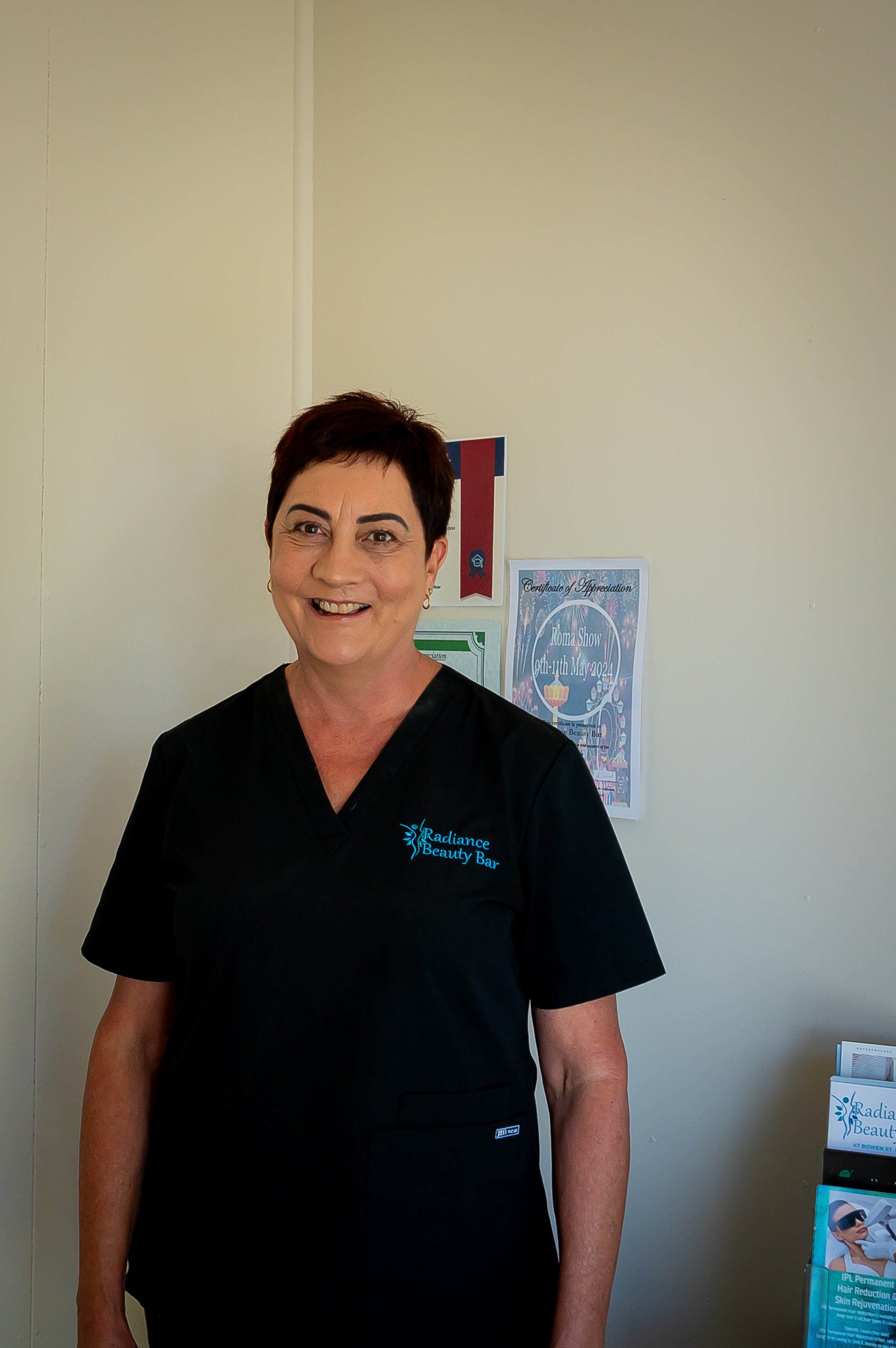 A woman with short dark hair, wearing a black medical uniform with the logo Radiance Beauty Bar, standing indoors in front of posters and brochures related to beauty treatments.