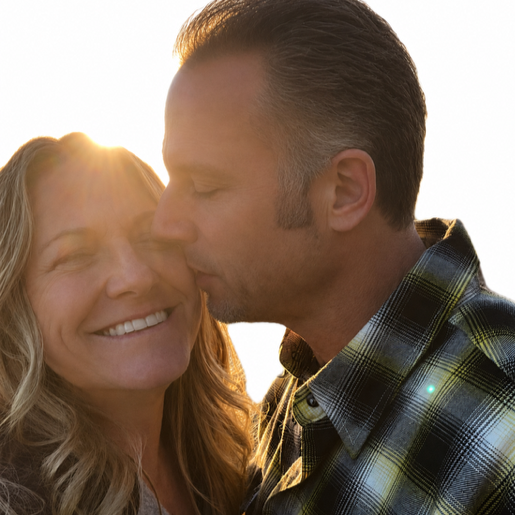 A man gently kisses a smiling woman with wavy hair on the forehead during sunset.