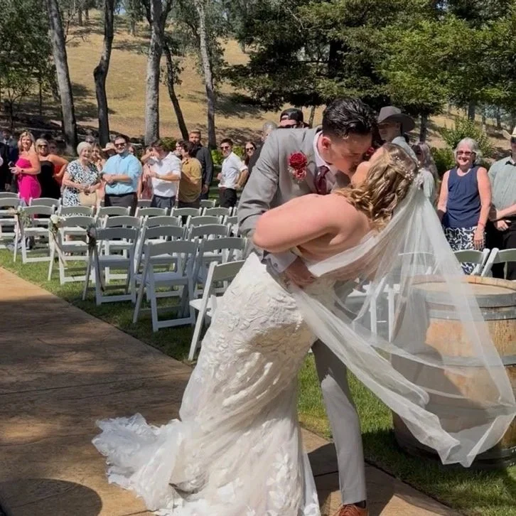 A bride and groom kiss during their outdoor wedding ceremony, with guests standing in the background on a sunny day surrounded by trees.