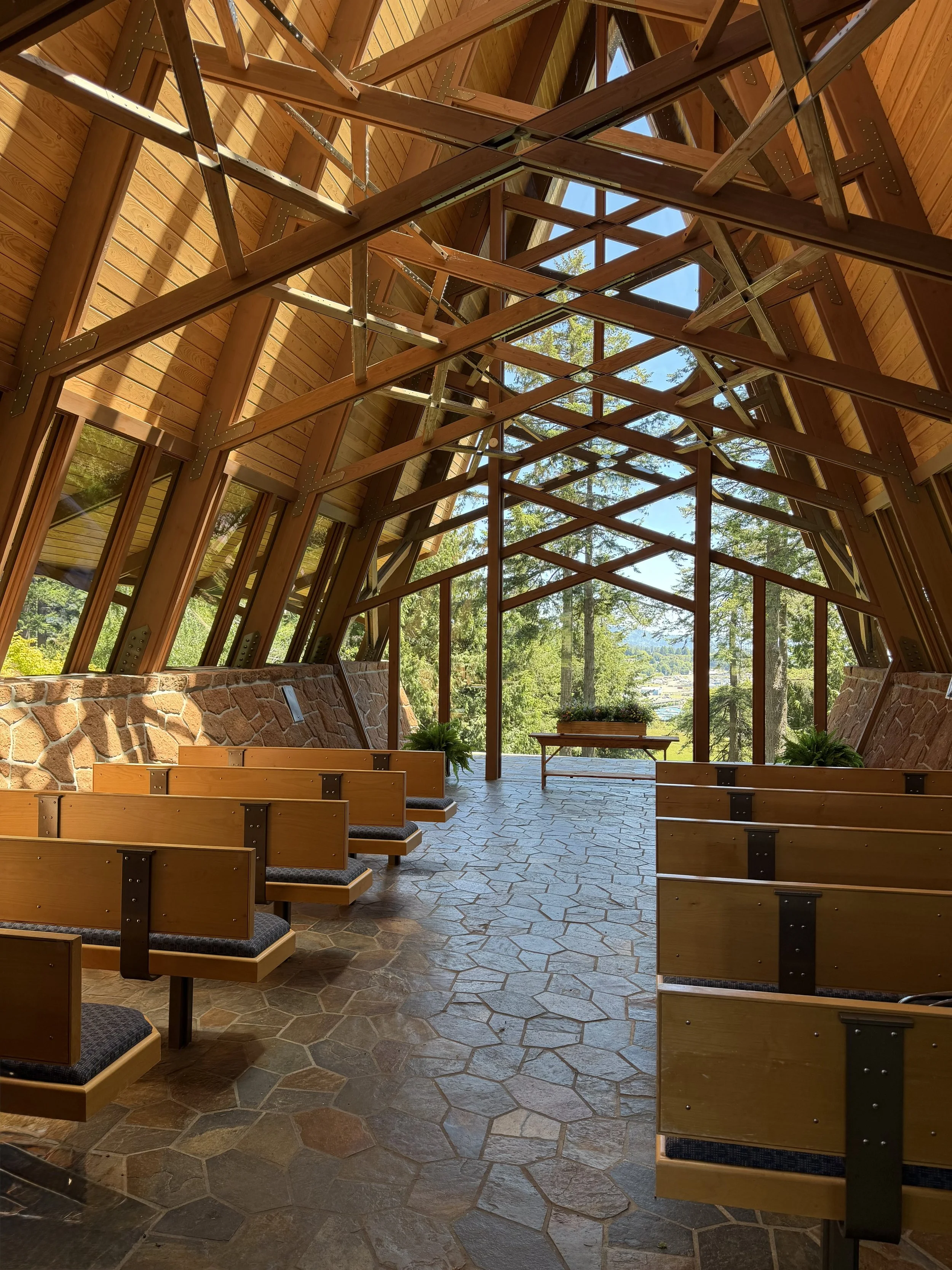 Inside view of a wooden chapel or church with large glass windows, wooden pews, stone flooring, and a view of trees and blue sky outside.