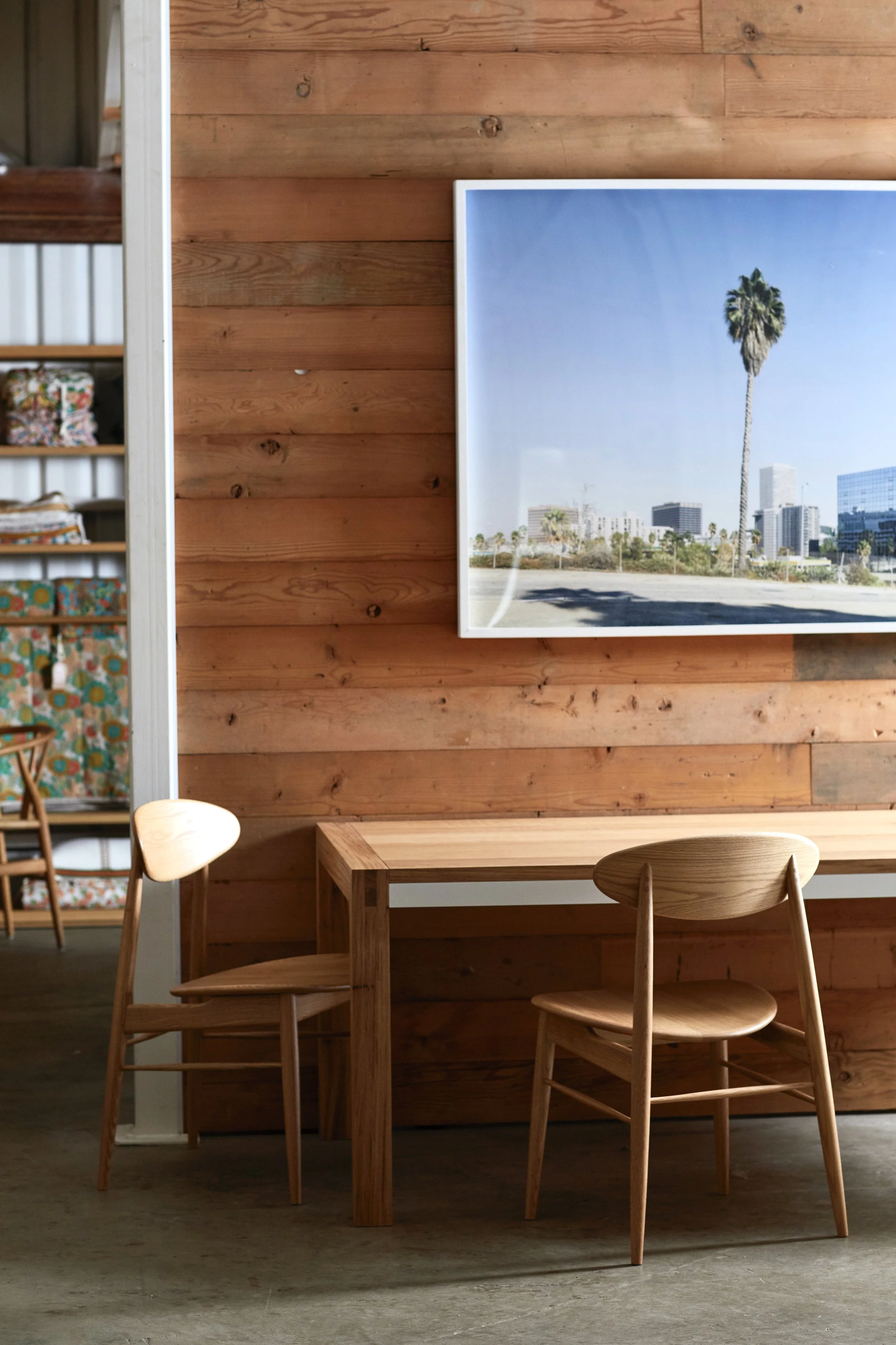 A wooden dining table with three matching wooden chairs in front of a wooden paneled wall. On the wall, there is a framed photo of a tall palm tree with a cityscape and clear blue sky in the background.