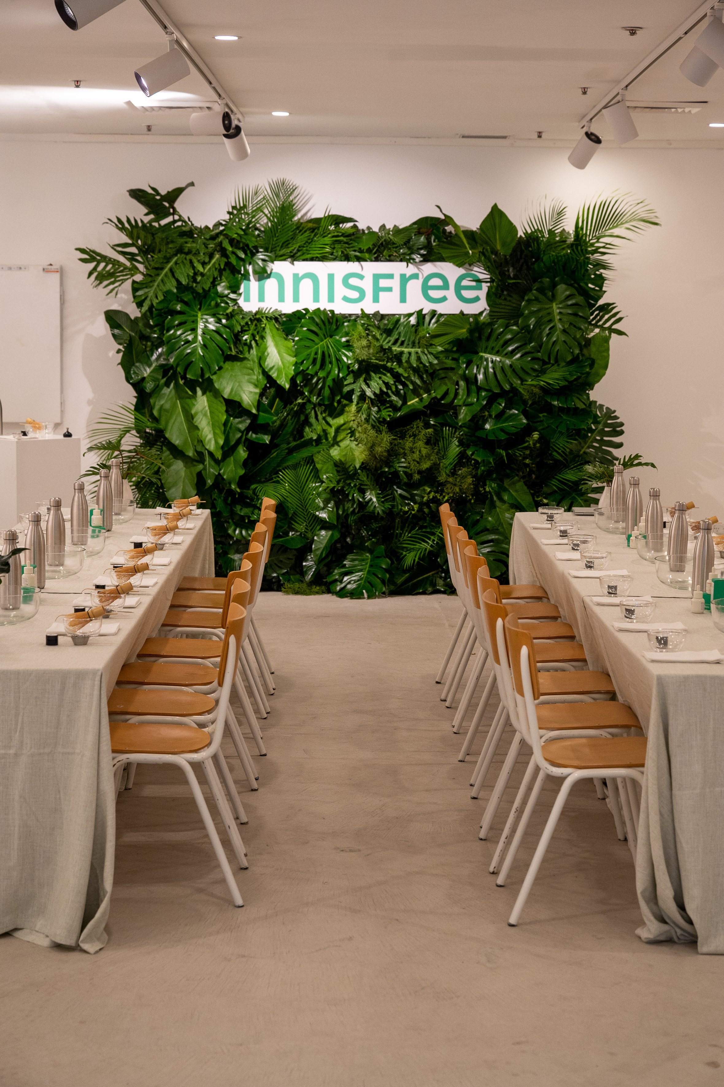 A conference room with two long tables covered in white tablecloths, arranged parallel with chairs on each side. The tables are set with glass water bottles, glasses, and small rolled-up napkins. Behind the tables, there is a large green wall made of