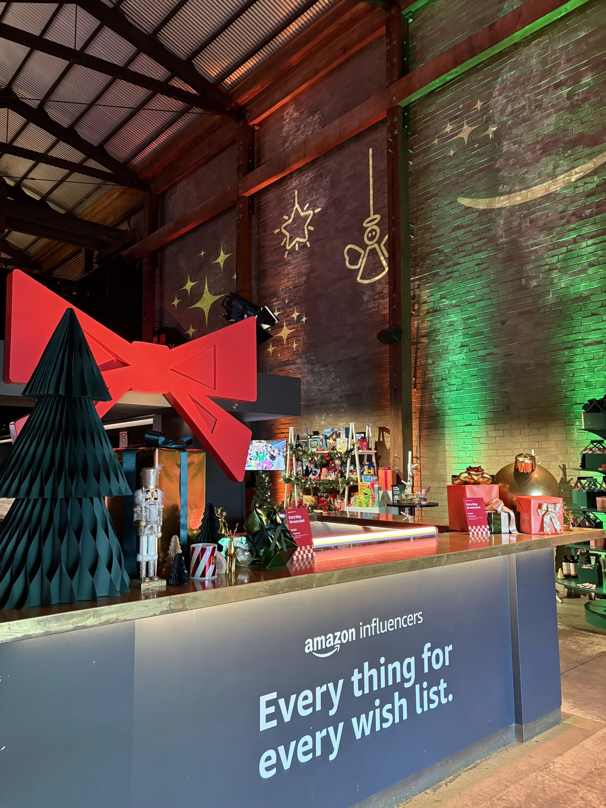 A store display area with Christmas decorations, including a red bow, gift boxes, and Christmas tree figures. The setting has a rustic, industrial style with exposed brick walls and metal beams, and is illuminated with festive lighting.