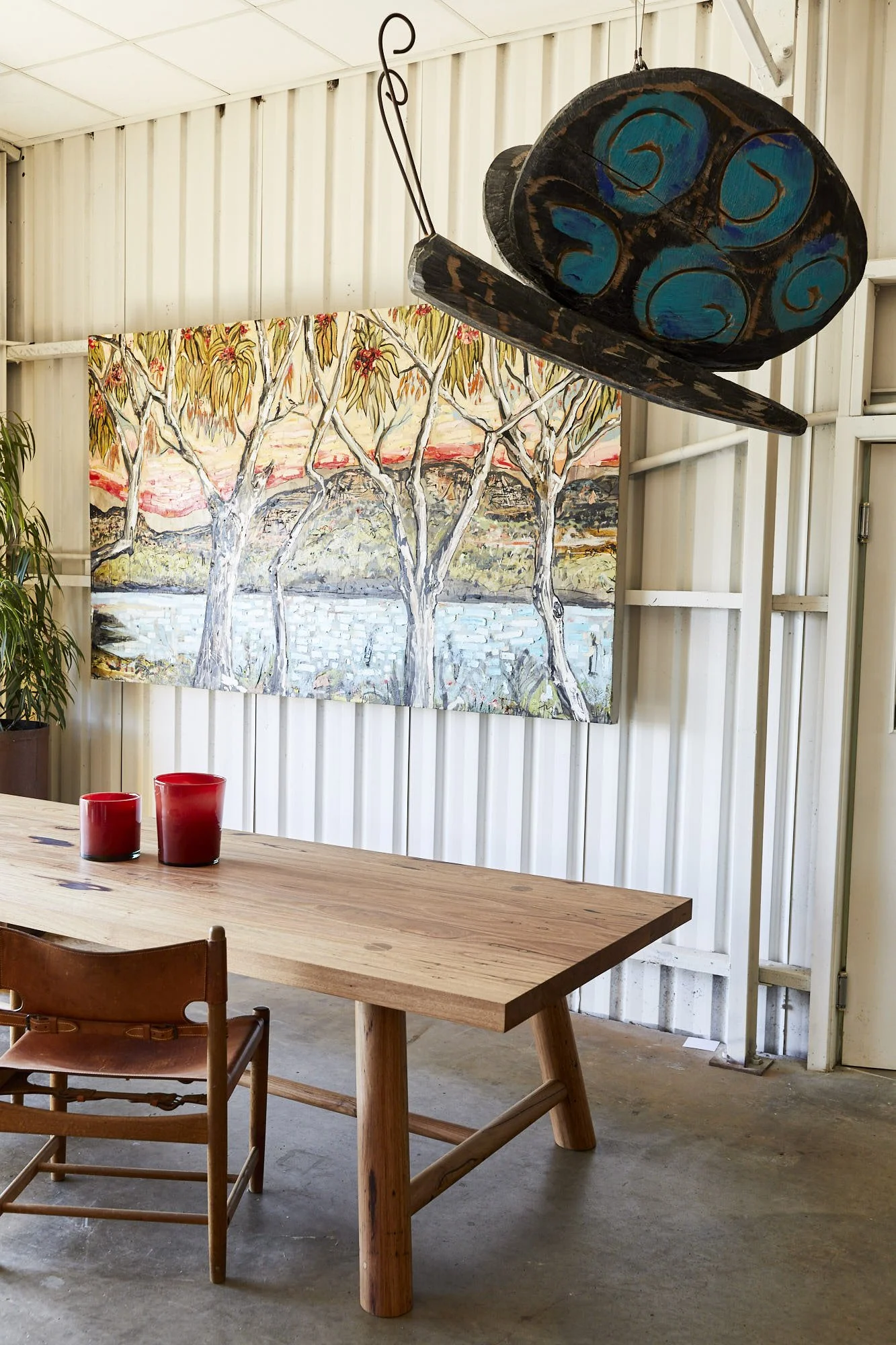 A large wooden table with two red candles on it, in a room with metal walls and a painting of trees and a sunset in the background, with a vintage wooden chair in the foreground and a large hanging wooden sculpture of a traditional canoe with blue pa