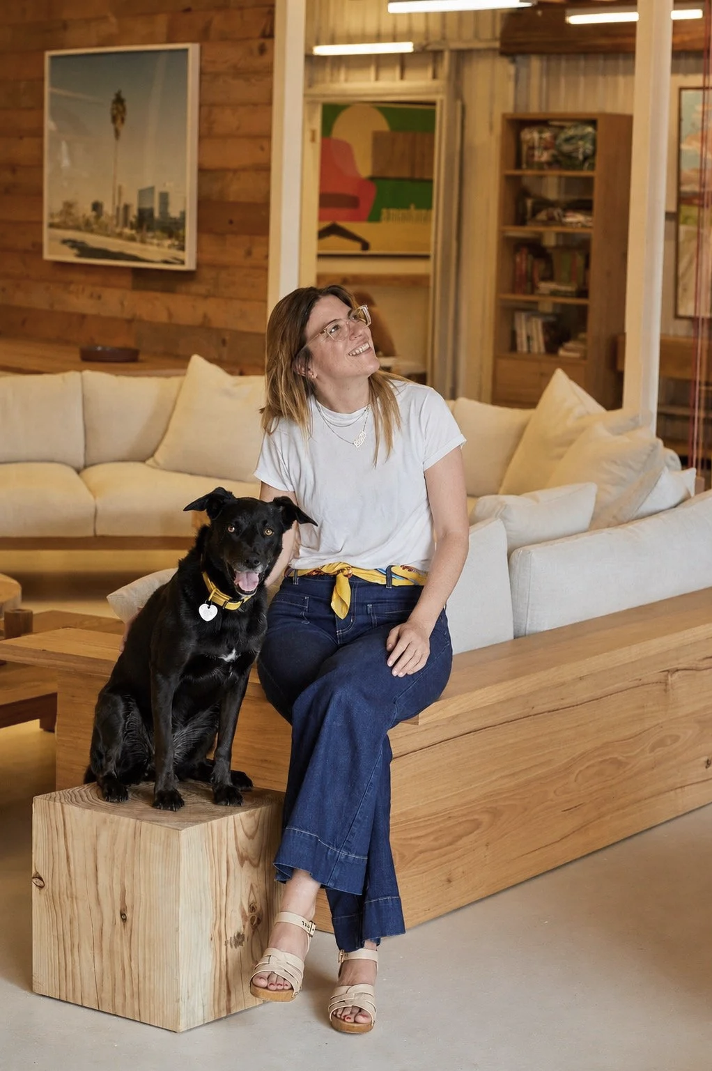 A woman sitting on a wooden bench with a black dog sitting beside her in a cozy living room with wood-paneled walls, artwork, and shelves filled with books.