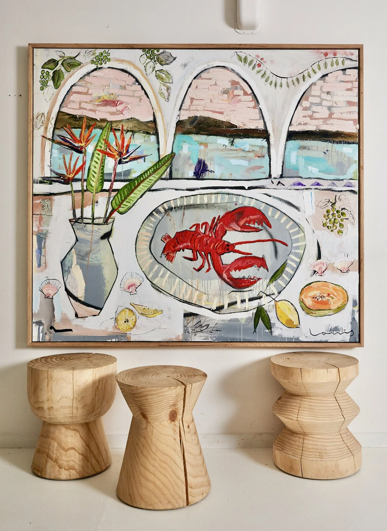 A colorful painting featuring a seaside landscape with arched windows, a flower vase with tropical flowers, a plate with a lobster, and lemon slices. The artwork is displayed on a white wall, with three wooden stools below.