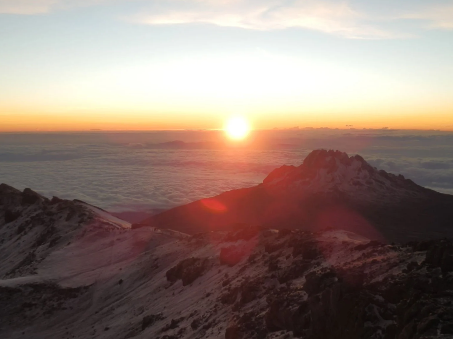 Sunrise over snow-capped Kilimanjaro's peaks with a sea of clouds below and a cloudy sky above.