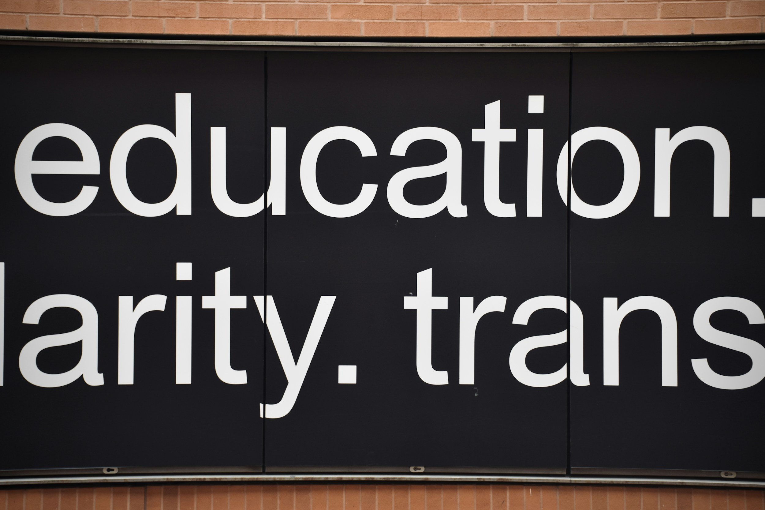 Close-up of a black sign with white text displaying the words 'education', 'art', 'trans'. The sign is mounted on a brick wall.