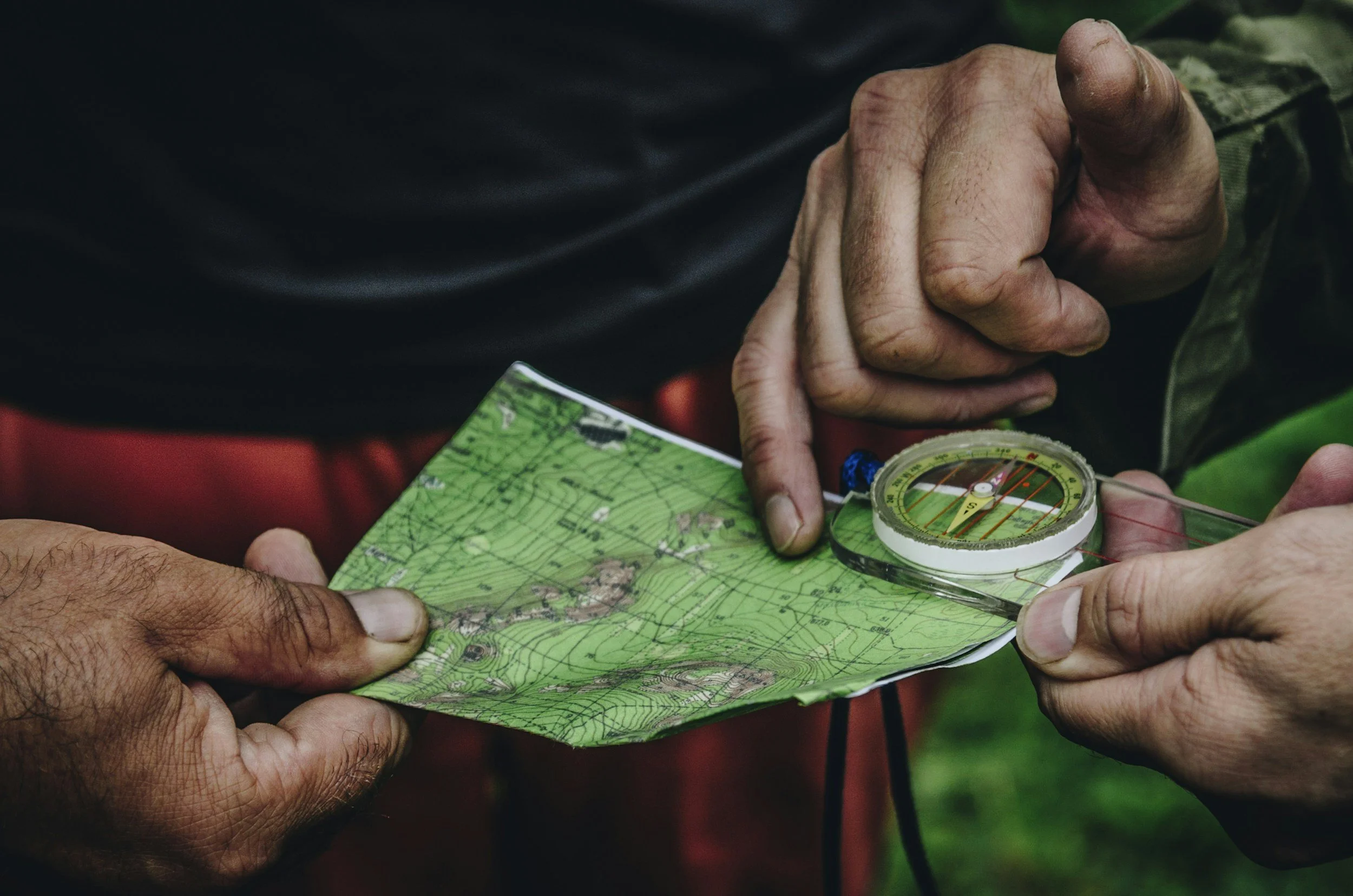 Several hands holding a topographic map, with a compass on top, suggesting outdoor navigation or hiking activity.