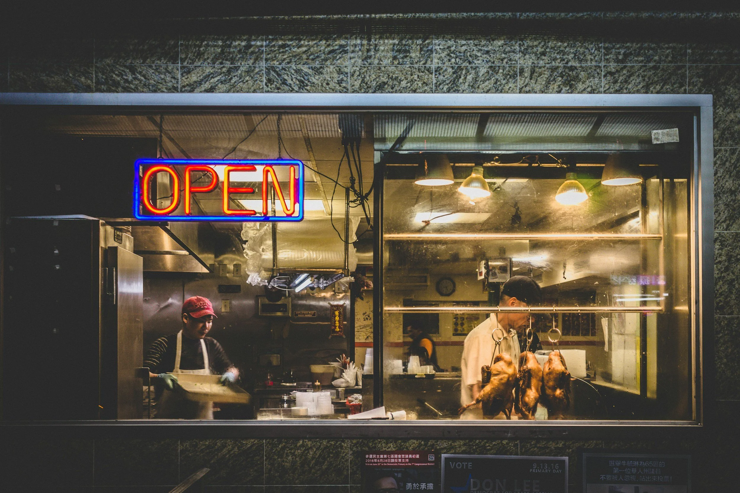 Nighttime view of a food stand or restaurant with a neon "OPEN" sign, through a large glass window showing workers preparing food inside, including hanging meats and kitchen equipment.