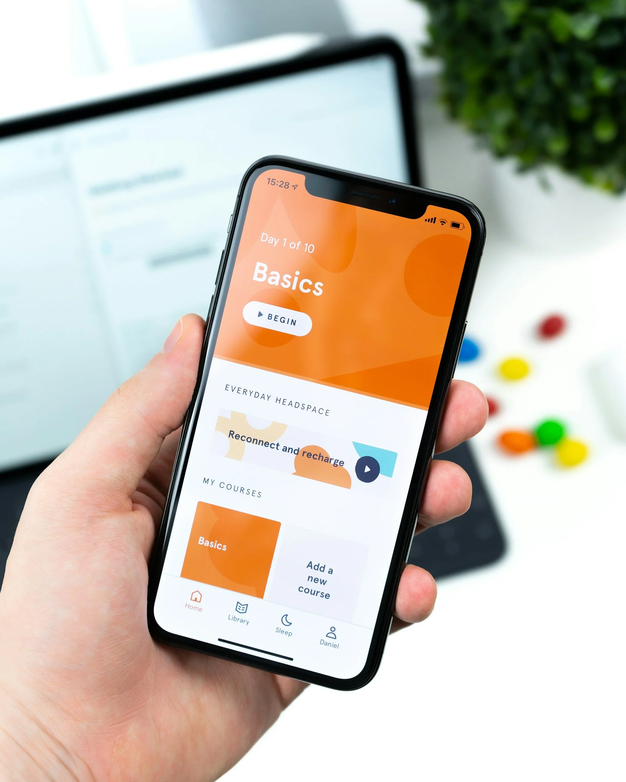 A person holding a smartphone displaying a health or fitness app with the title 'Basics' on a bright orange background, with a white button labeled 'Begin'. The background shows a blurred workspace or study area with a computer monitor and a green plant.